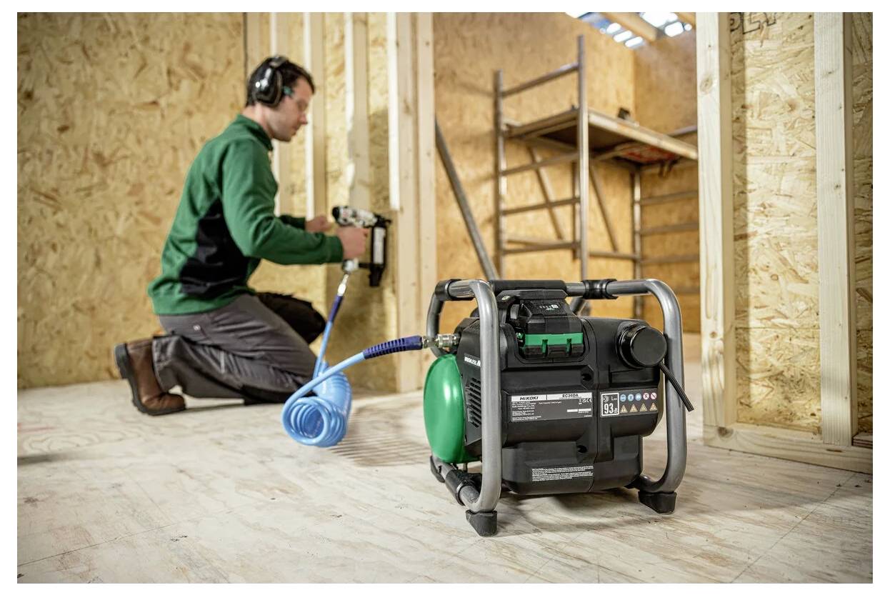 Eine Person mit Gehörschutz verwendet eine Druckluftnagelmaschine, die an einen tragbaren Kompressor angeschlossen ist, in einer Holzbau-Umgebung.