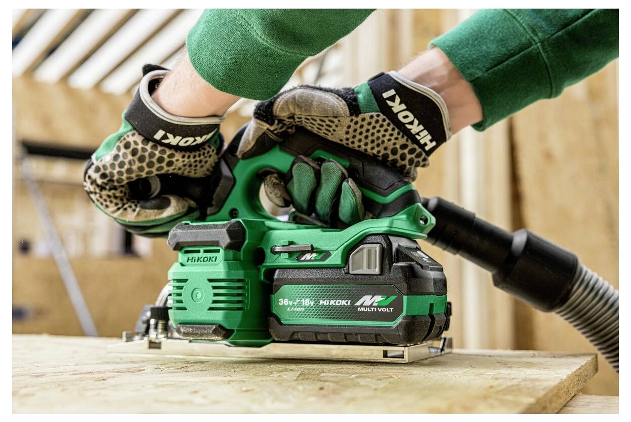 Person mit Handschuhen, die ein grün-schwarzes tragbares Elektrowerkzeug auf einer Holzoberfläche in einer Werkstatt benutzt.