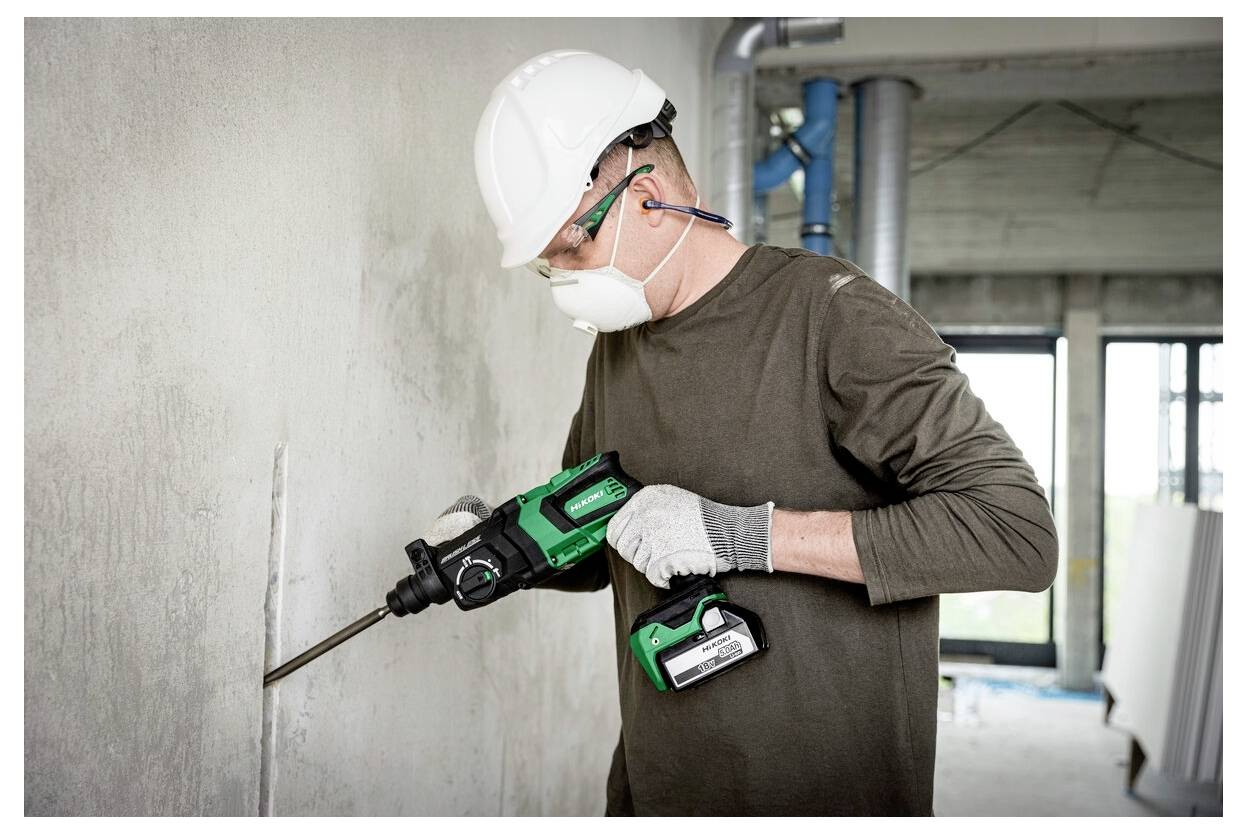 Ein Arbeiter mit Sicherheitsausrüstung verwendet ein grünes Elektrowerkzeug, um eine Betonwand in einem teilweise fertiggestellten Gebäude zu meißeln.