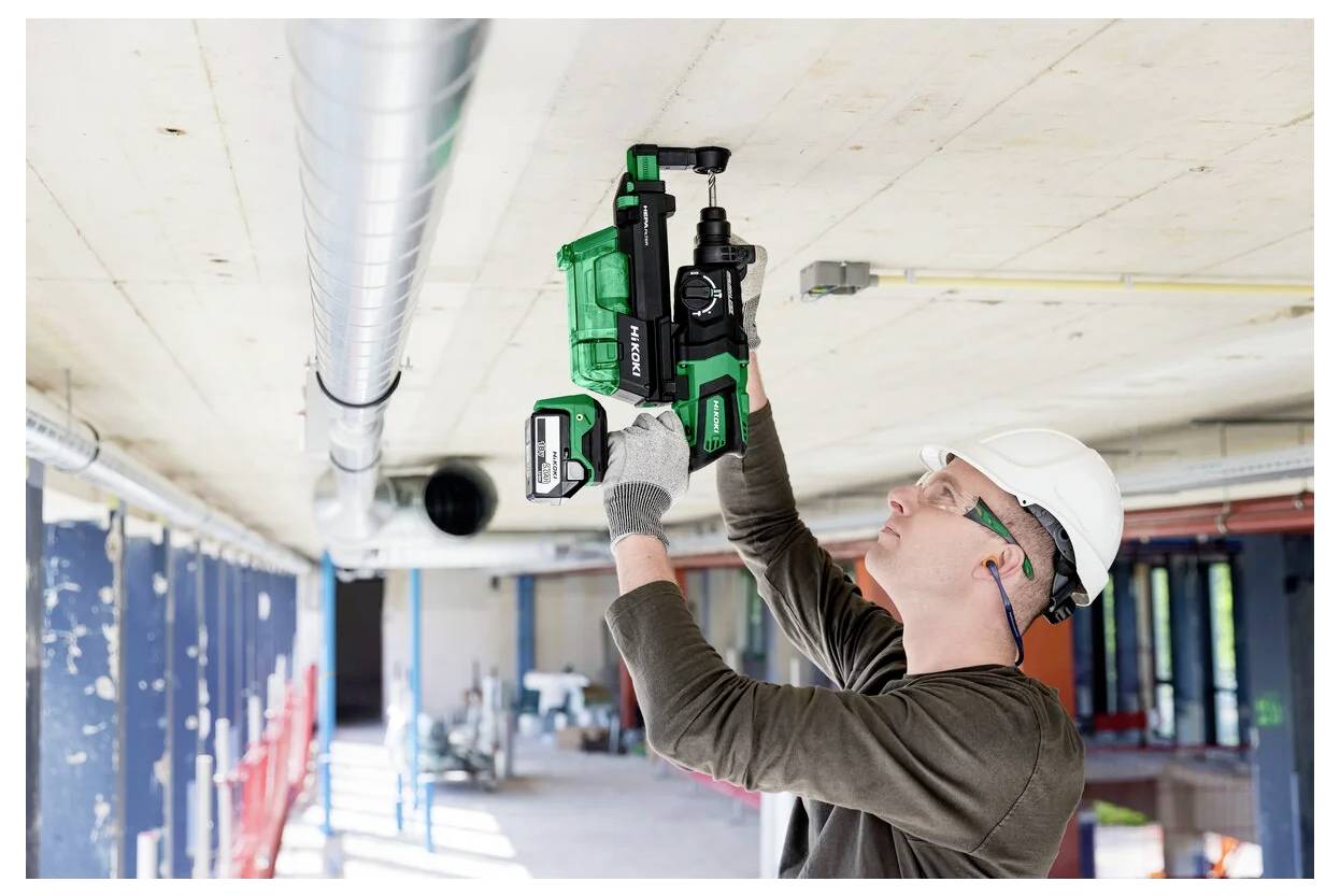 Ein Bauarbeiter in Schutzausrüstung verwendet ein grün-schwarzes Elektrowerkzeug, um an der Decke eines großen, hell erleuchteten Industrieraums zu arbeiten.
