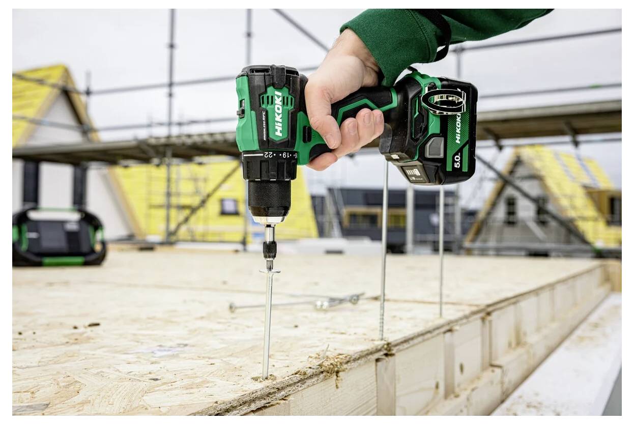 Eine Hand bedient einen grünen kabellosen Bohrer, der eine Schraube in ein Holzbrett auf einer Baustelle einschraubt, während im Hintergrund Gerüste und Gebäude zu sehen sind.