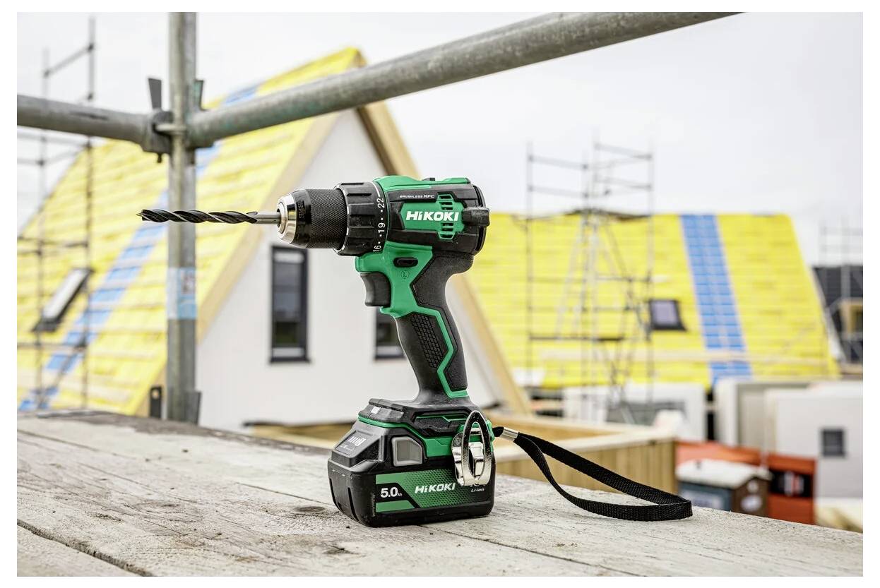 Eine kabellose Bohrmaschine mit schwarzem und grünem Design ruht auf einem Holzbrett an einer Baustelle, mit teilweise fertiggestellten Häusern im Hintergrund.