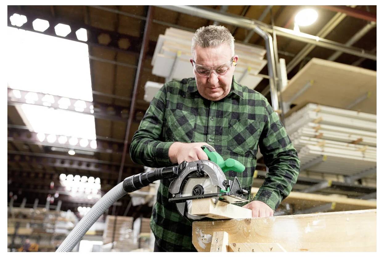 Eine Person mit Sicherheitsbrille bedient eine Kreissäge auf einem Holzbrett in einer Werkstatt mit Regalen voller Materialien im Hintergrund.