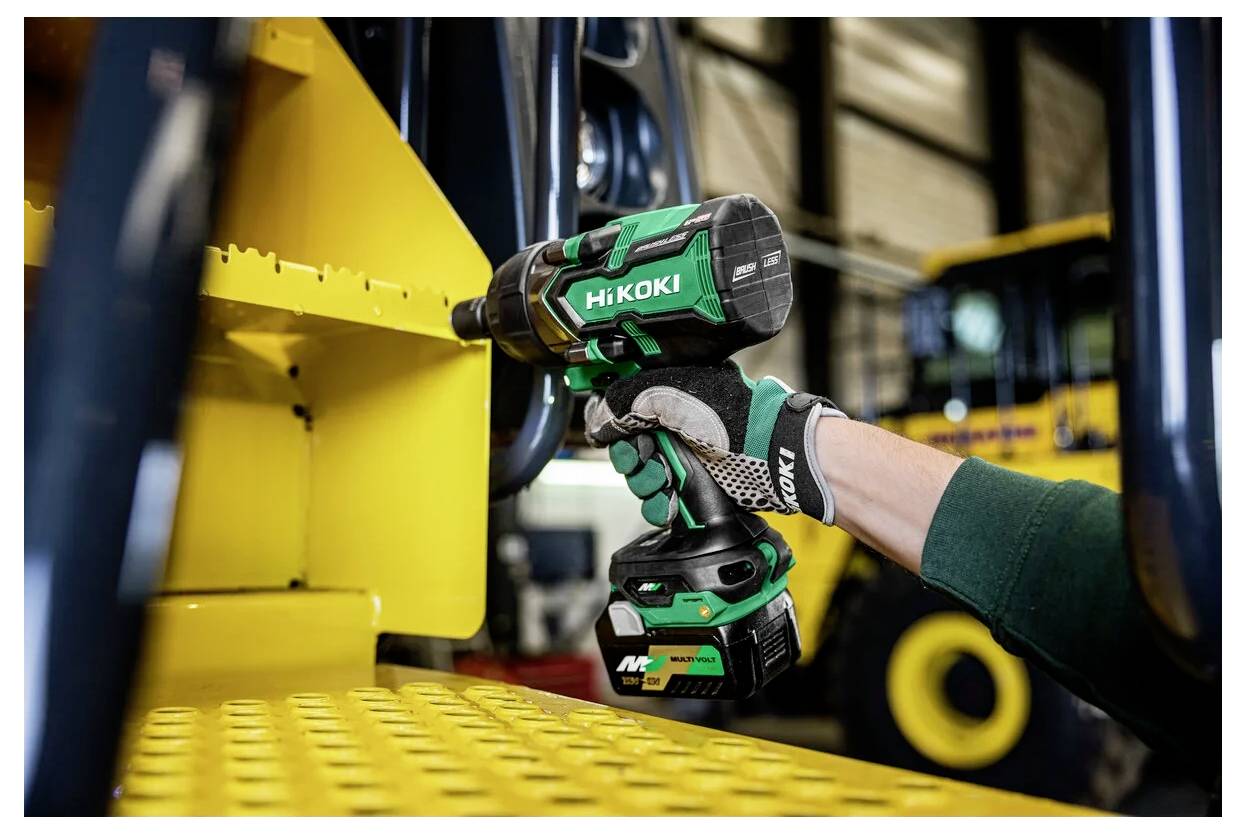 Eine Hand in einem Handschuh benutzt einen grünen kabellosen Schlagschrauber, um eine Schraube an gelbem Industriegerät in einer Werkstattumgebung anzuziehen.