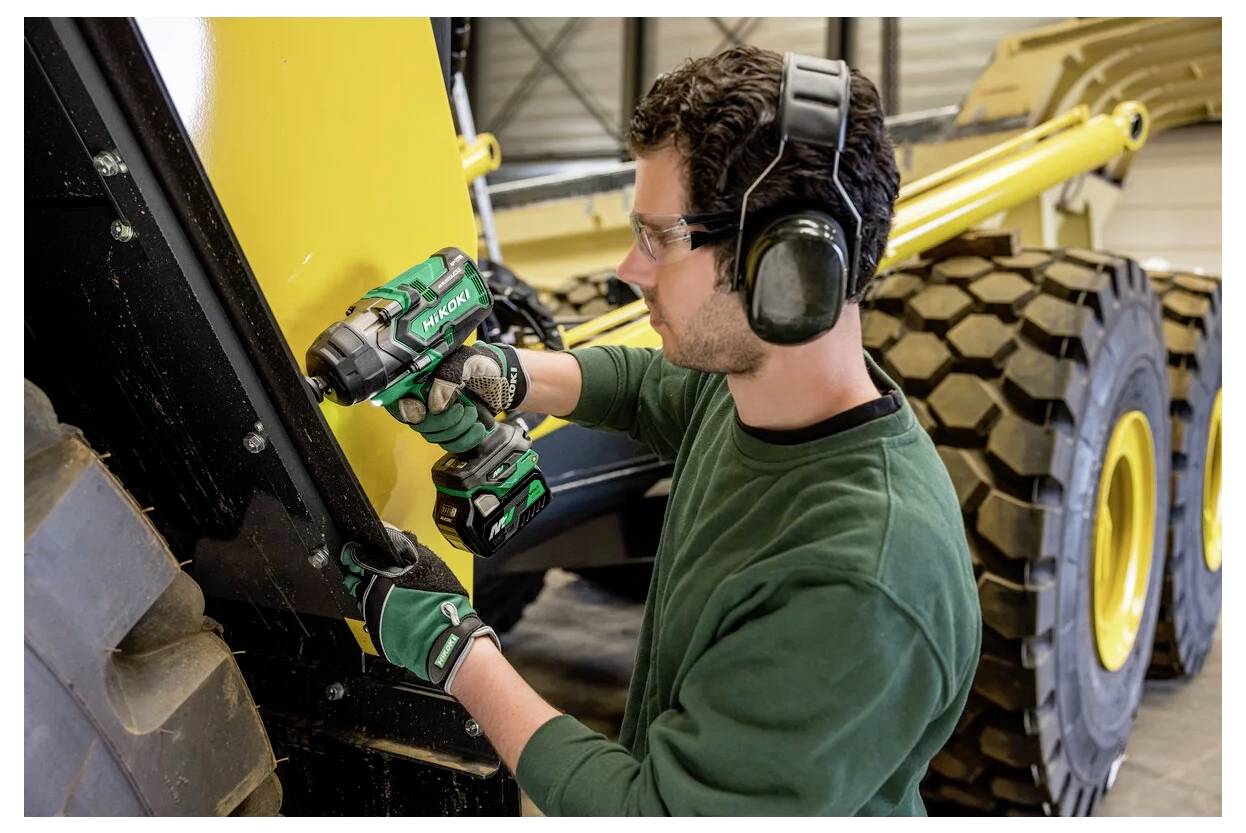 Ein Mann mit Sicherheitsausrüstung bedient eine Bohrmaschine an einer Maschine. Er ist konzentriert bei seiner Arbeit, im Hintergrund sind große Fahrzeugteile zu sehen.