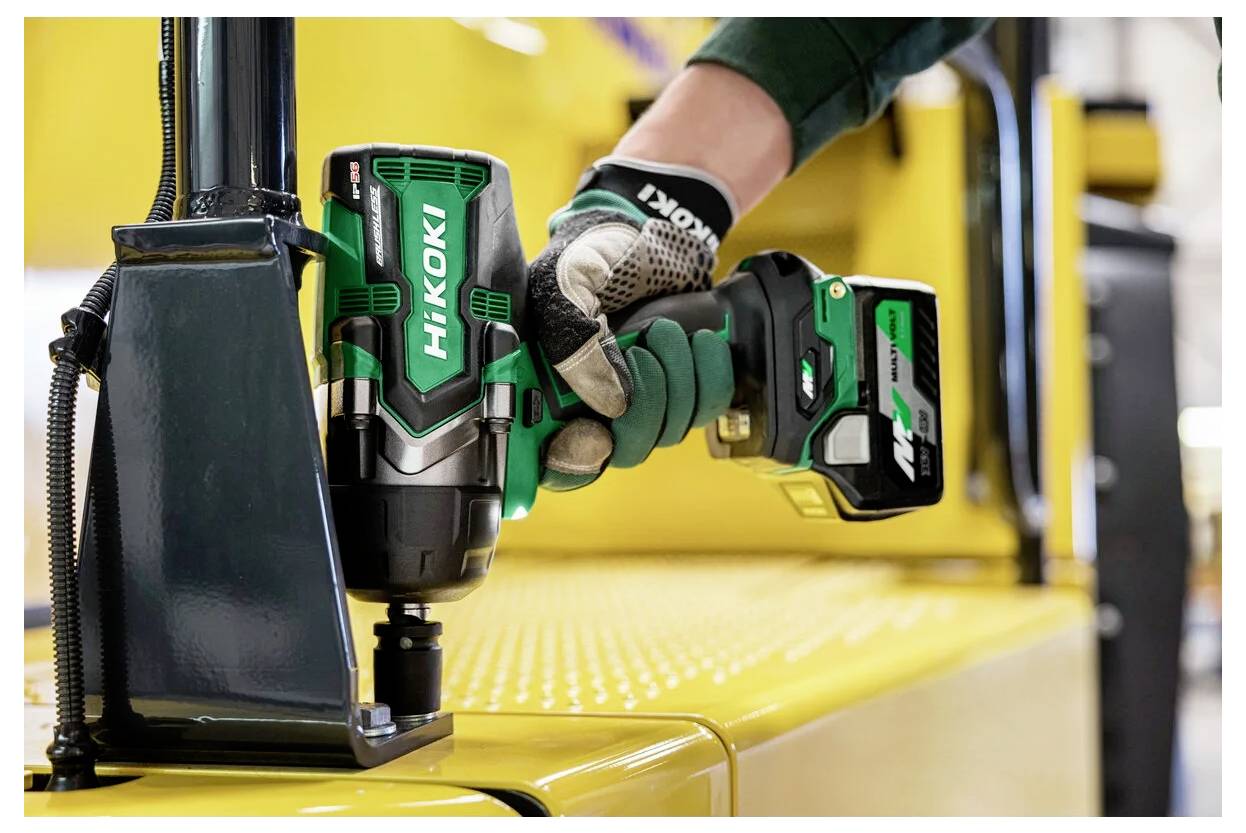 Eine Person, die einen grünen kabellosen Akkuschrauber auf einer gelben Metalloberfläche im Innenraum benutzt. Der Hintergrund zeigt industrielle Ausrüstung.