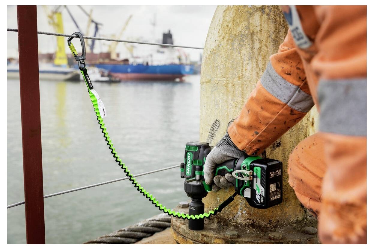 Ein Arbeiter in orangefarbener Sicherheitsausrüstung benutzt einen Akkuschrauber an einer Metallstruktur in der Nähe eines Hafens, mit Frachtchiffen und Kränen im Hintergrund.