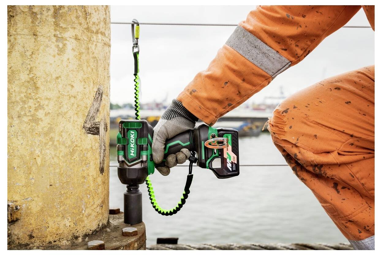 Eine Person trägt einen orangefarbenen Sicherheitsanzug und Handschuhe und verwendet ein grünes HiKOKI-Elektrowerkzeug, um an einer großen Metallstruktur am Wasser zu arbeiten.