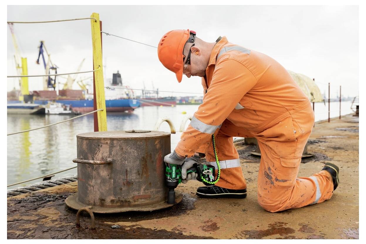 Ein Arbeiter in orangefarbener Schutzkleidung kniet auf einem Dock und verwendet eine Bohrmaschine an einem Metallgegenstand. Im Hintergrund sind große Schiffe und Kräne zu sehen.