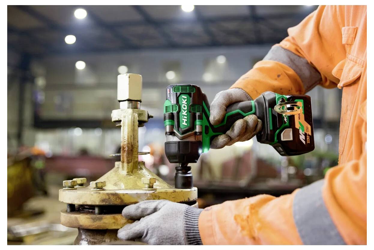 Eine Person in einer orangefarbenen Arbeitsjacke verwendet ein HiKOKI-Elektrowerkzeug, um einen Bolzen an industrieller Ausrüstung in einer Werkstatt zu befestigen.
