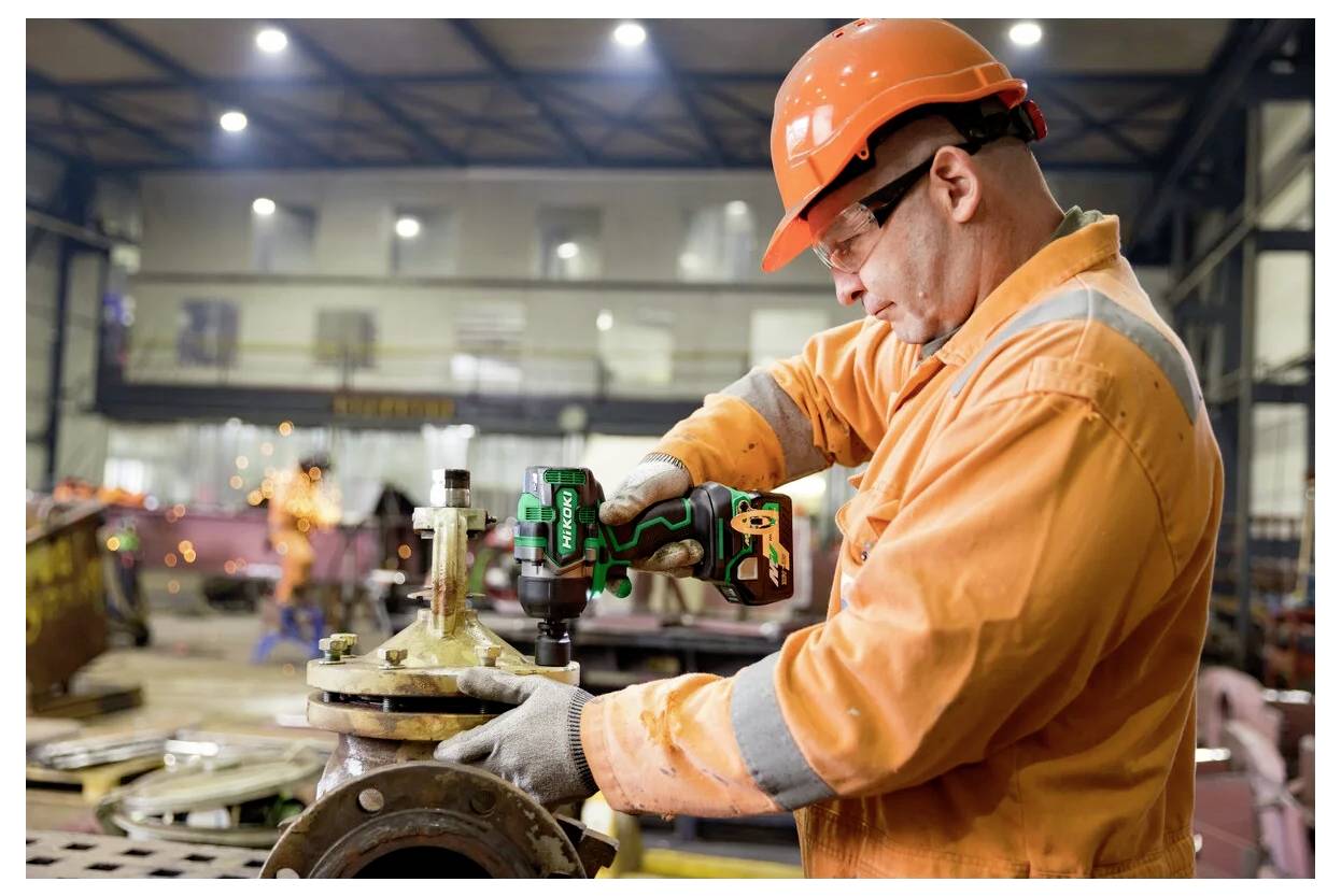 Ein Arbeiter in einem orangefarbenen Sicherheitsanzug und Helm bedient eine Bohrmaschine an industrieller Maschinenausrüstung in einer Fabrikhalle, konzentriert und präzise.