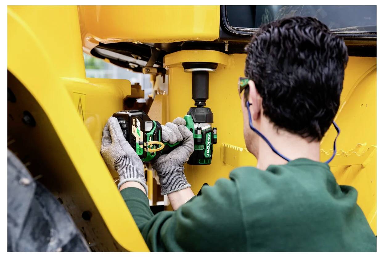 Eine Person mit Handschuhen und Gehörschutz verwendet einen grünen kabellose Bohrschrauber, um an einer großen gelben Maschine zu arbeiten, möglicherweise für Wartungszwecke.