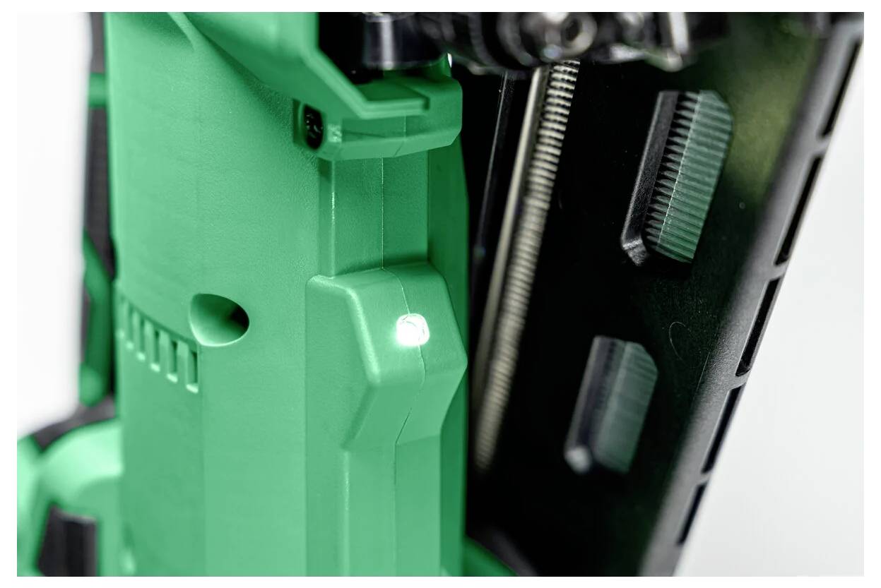 Eine Nahaufnahme eines grünen Werkzeugs, möglicherweise ein Elektrowerkzeug oder eine Nagelpistole, mit sichtbarer LED-Leuchte, die anzeigt, dass es in Betrieb ist. Details der Metallteile sind zu erkennen.