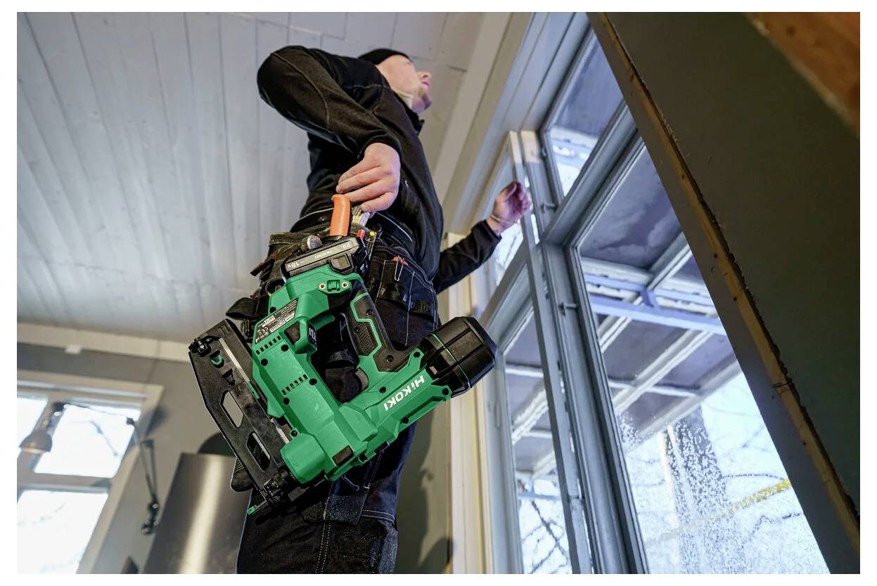 Ein Arbeiter mit einer kabellosen Bohrmaschine steht neben einem Fenster, trägt einen Werkzeuggürtel und scheint sich mitten in einer Bauaufgabe im Innenbereich zu befinden.