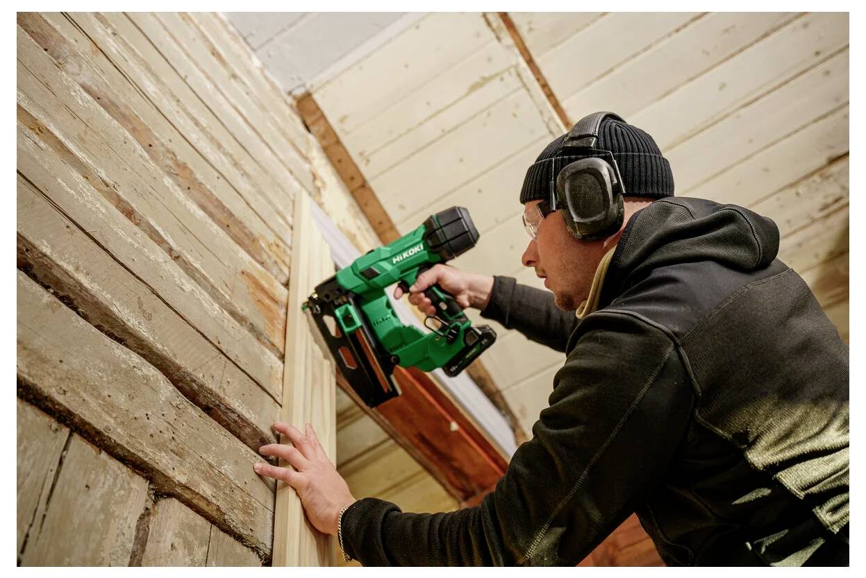 Eine Person in Schutzausrüstung verwendet eine Nagelpistole, um Holzplatten an einer Wand zu befestigen, was auf Bauarbeiten im Gange hinweist.