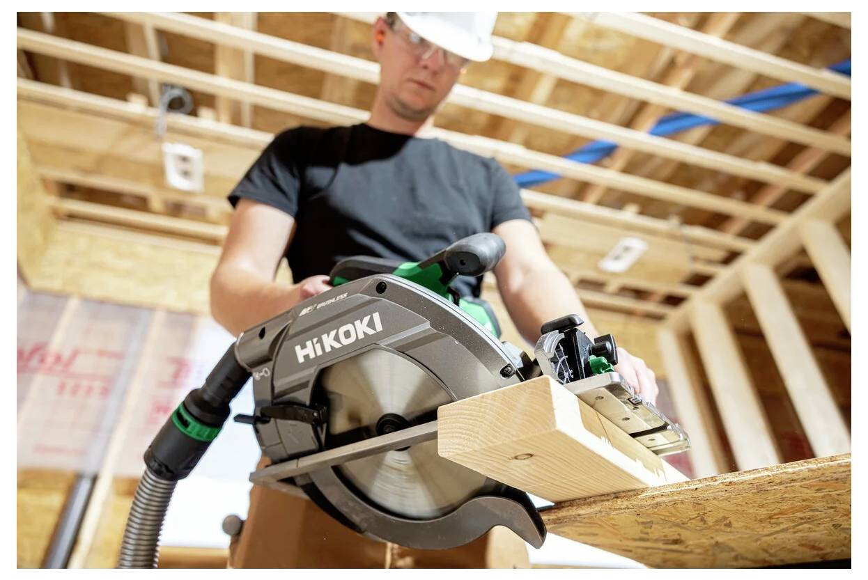 Ein Bauarbeiter mit Schutzhelm verwendet eine HiKOKI-Kreissäge, um einen Holzbalken in einem teilweise fertiggestellten Gebäude zu sägen.