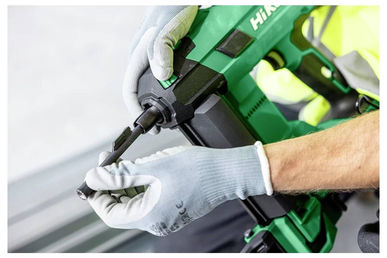 Eine Person mit Handschuhen bedient ein grünes Elektrowerkzeug und führt eine Komponente an dessen Ende ein, möglicherweise für Bauarbeiten.