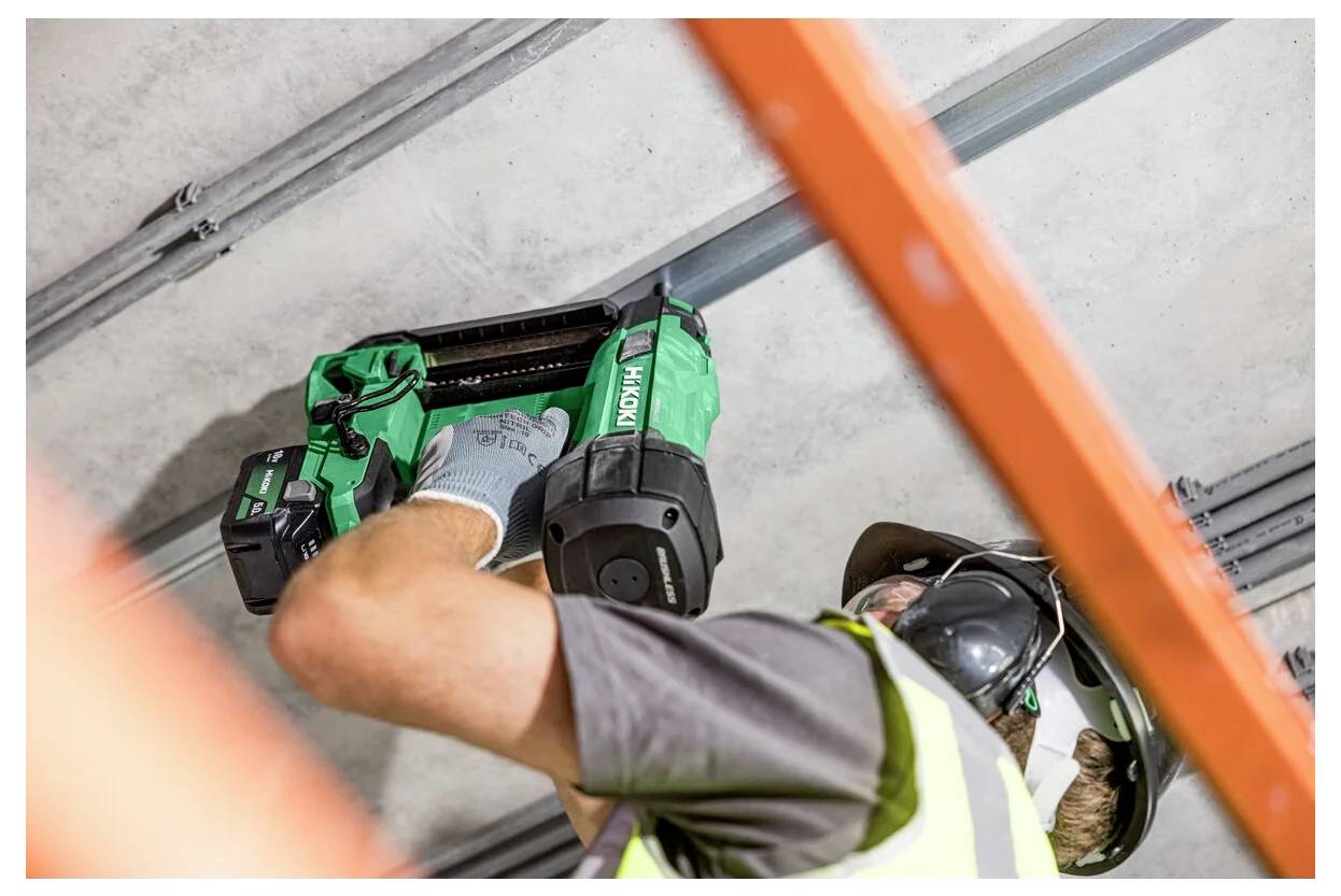 Ein Bauarbeiter mit Schutzausrüstung verwendet ein grünes Elektrowerkzeug, um Kabel an einer Decke in einer industriellen Umgebung zu befestigen.