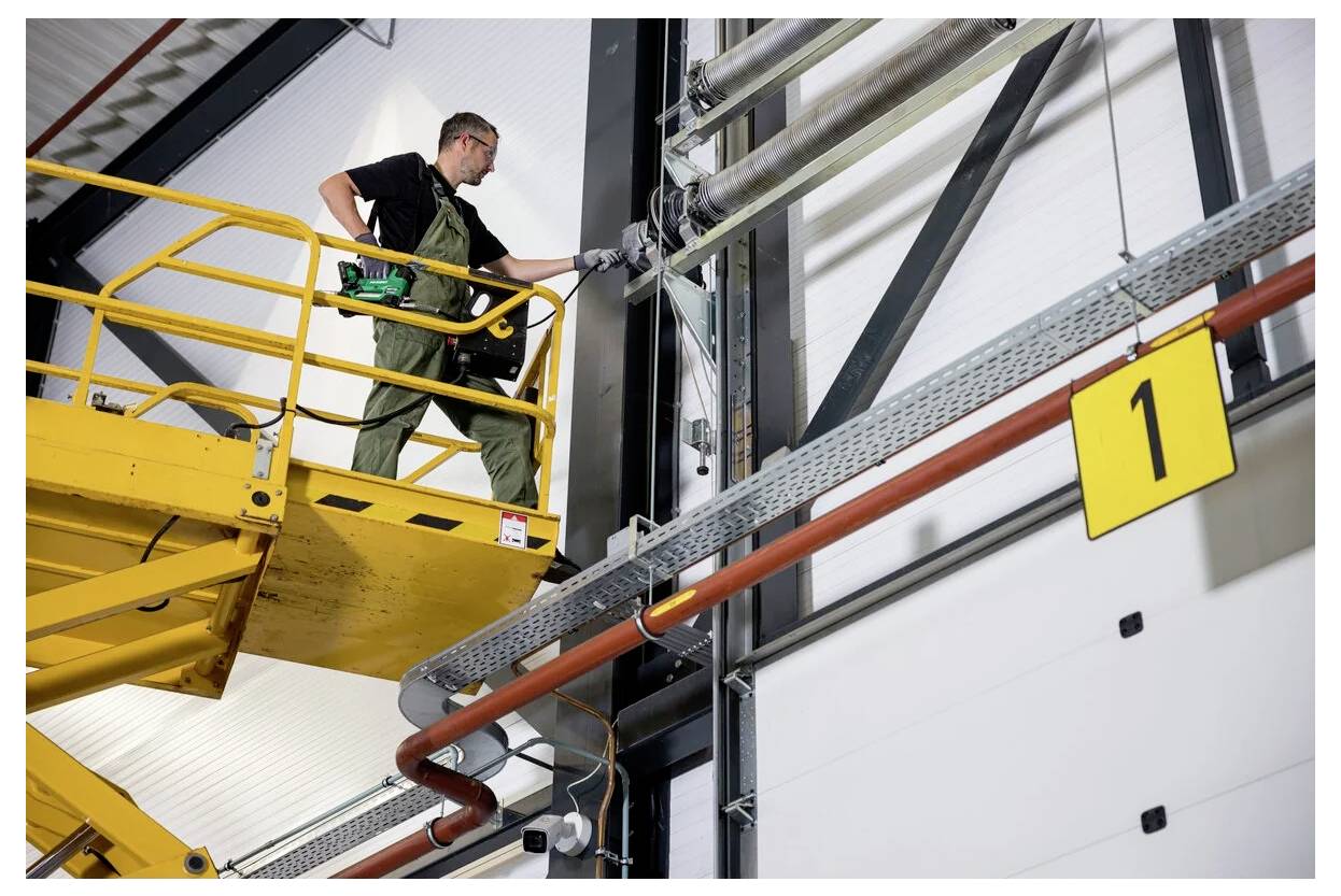 Arbeiter in Schutzausrüstung auf einem gelben Hubsteiger, der Wartungsarbeiten an Rohren in einer industriellen Umgebung durchführt, mit einem nummerierten Schild in der Nähe.
