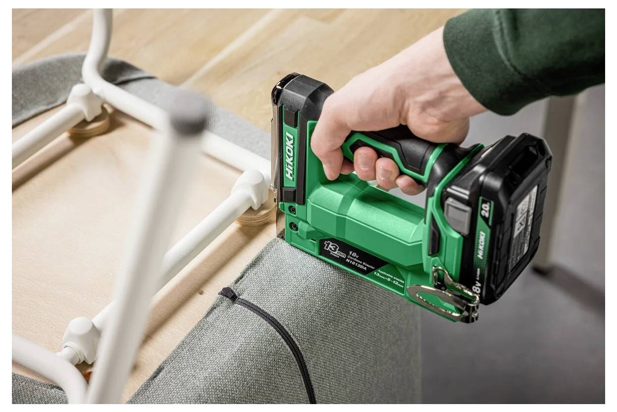 Eine Person verwendet eine grüne Tacker-Pistole, um Stoff am Rahmen eines Holzstuhls zu befestigen. Das Werkzeug ist nahe der Unterseite des Sitzes positioniert.