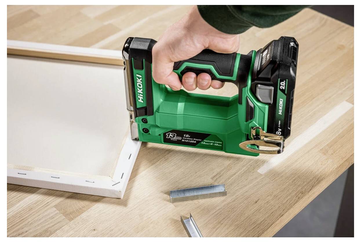 Eine Person verwendet eine grüne Nagelpistole, um einen Holzrahmen zu befestigen, wobei metallene Nägel auf dem Tisch sichtbar sind.