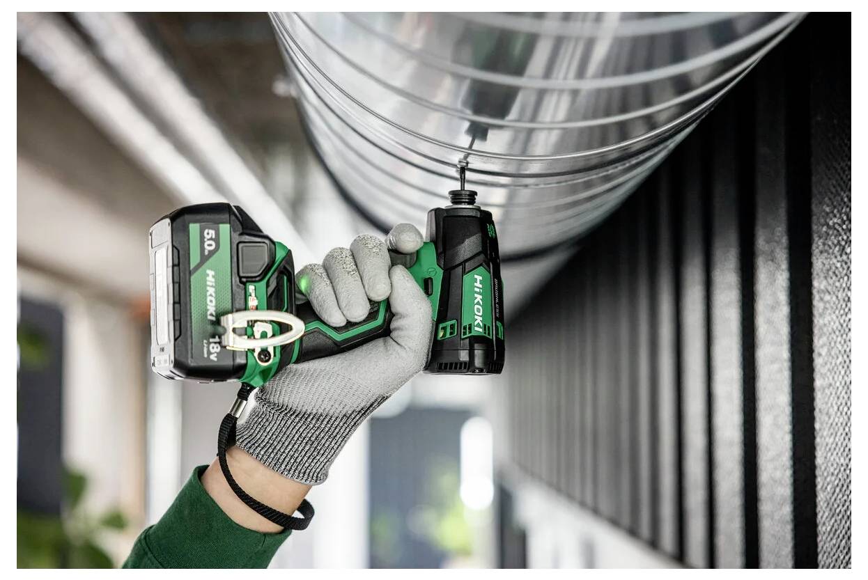 Eine Person mit Handschuhen verwendet einen grünen kabellosen Akkuschrauber, um eine Schraube in einen Metalllüftungskanal in einer industriellen Umgebung zu befestigen.