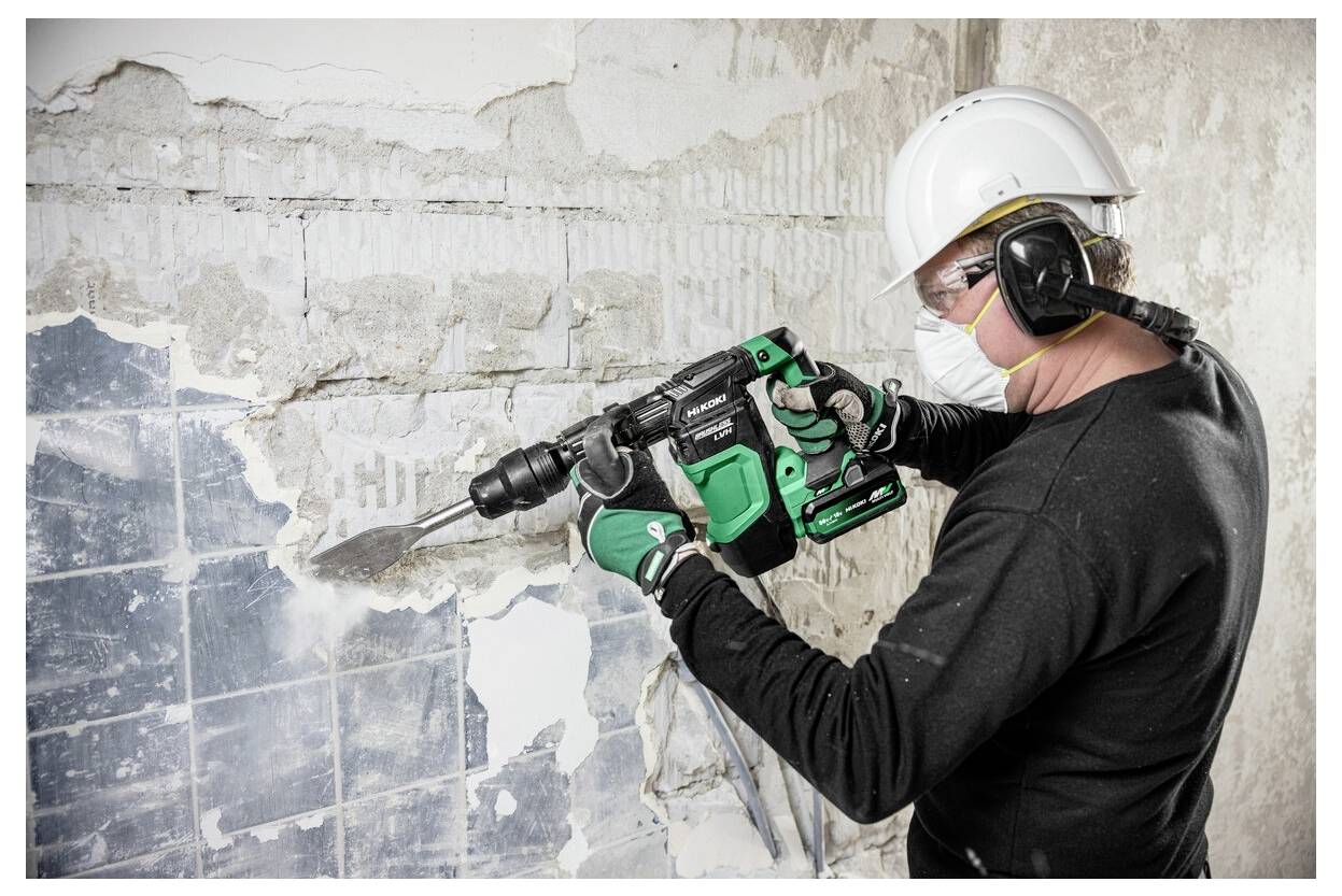Eine Person in Sicherheitsausrüstung verwendet ein grünes Elektrowerkzeug, um alte Fliesen von einer Wand zu entfernen, und trägt dabei einen Helm, Schutzbrille und eine Maske zum Schutz.