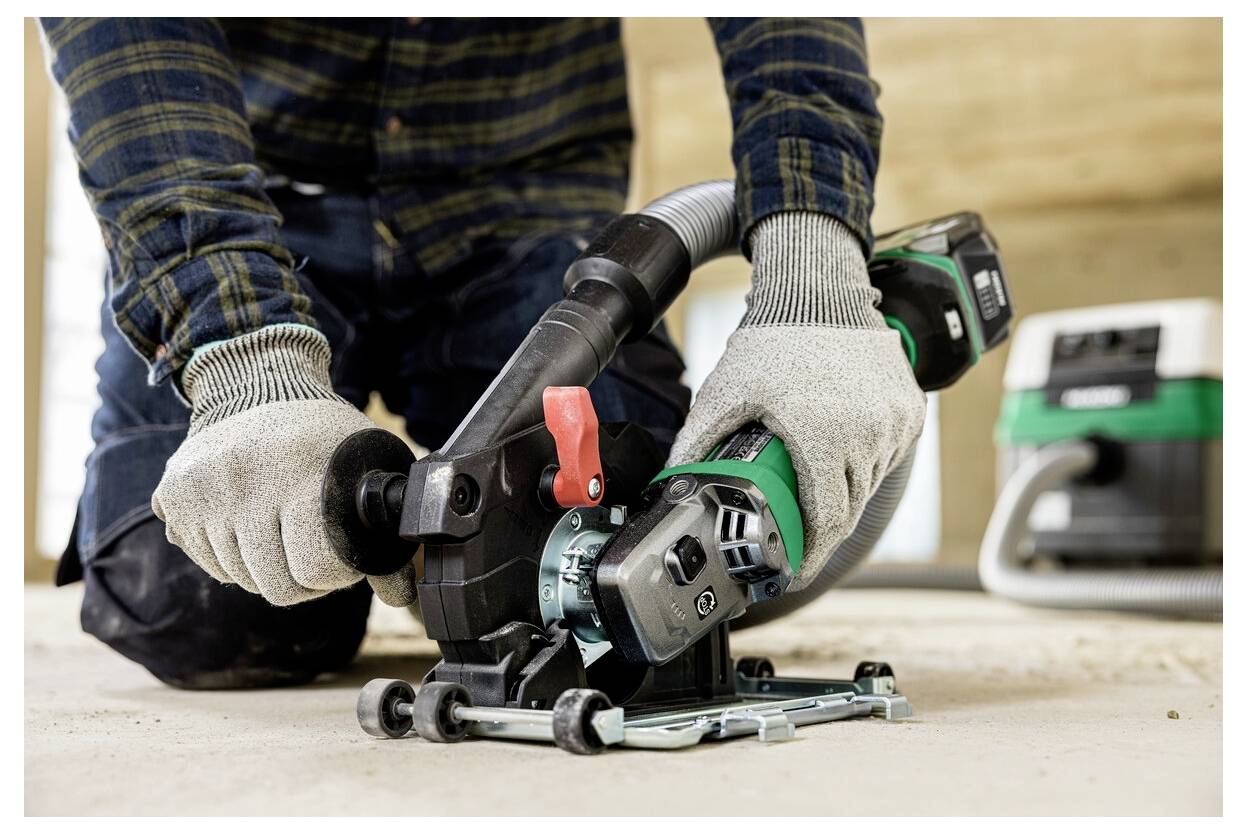 Eine Person kniet während der Verwendung eines tragbaren Elektrowerkzeugs zum Schneiden einer Oberfläche und trägt Schutzhandschuhe. Ein Absaugsystem ist zur Staubkontrolle angeschlossen.
