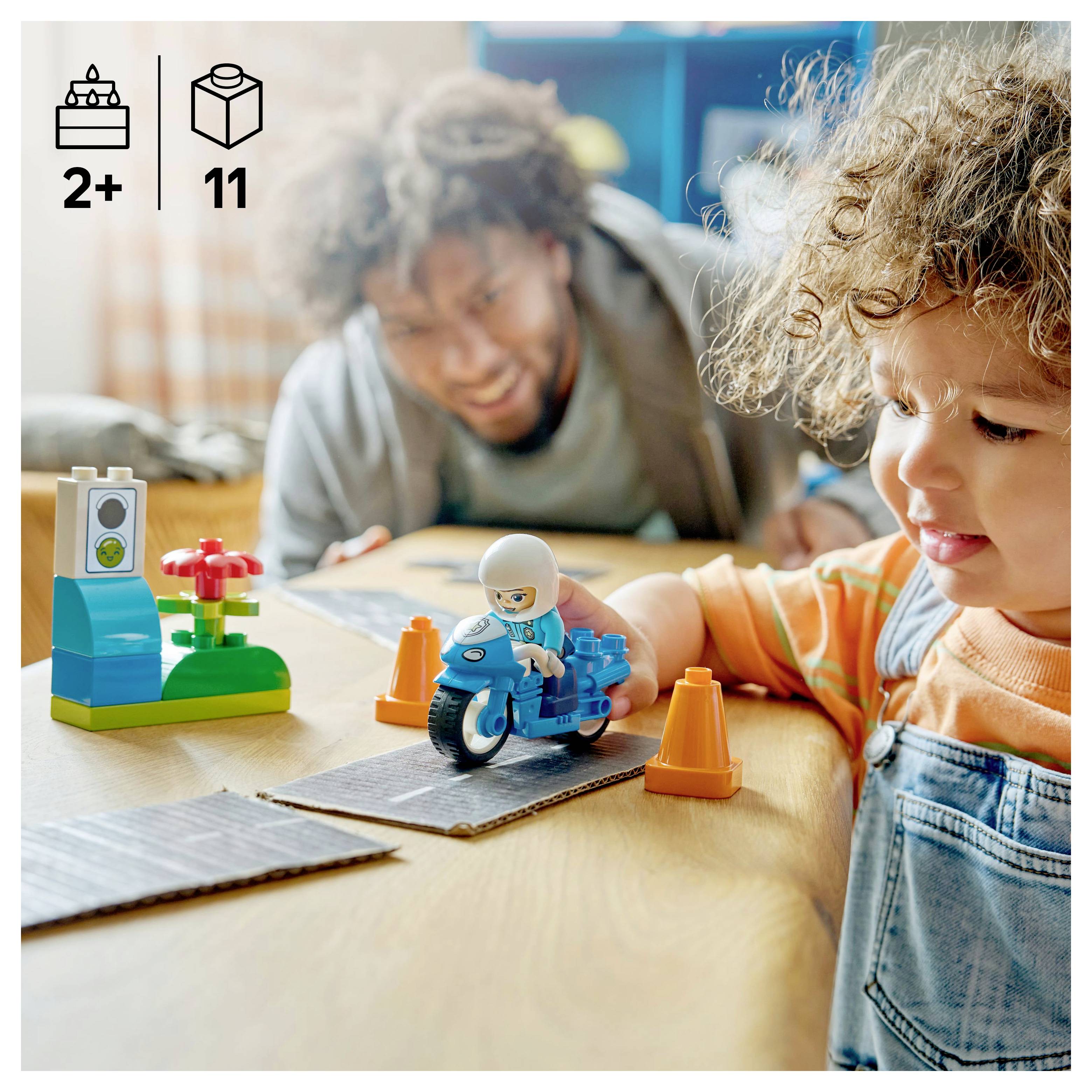 Kind, das mit einem Spielzeug-Motorrad auf einer Straßenbahn spielt, während ein Erwachsener im Hintergrund steht. Spielblöcke und Blumen liegen auf einem Tisch.