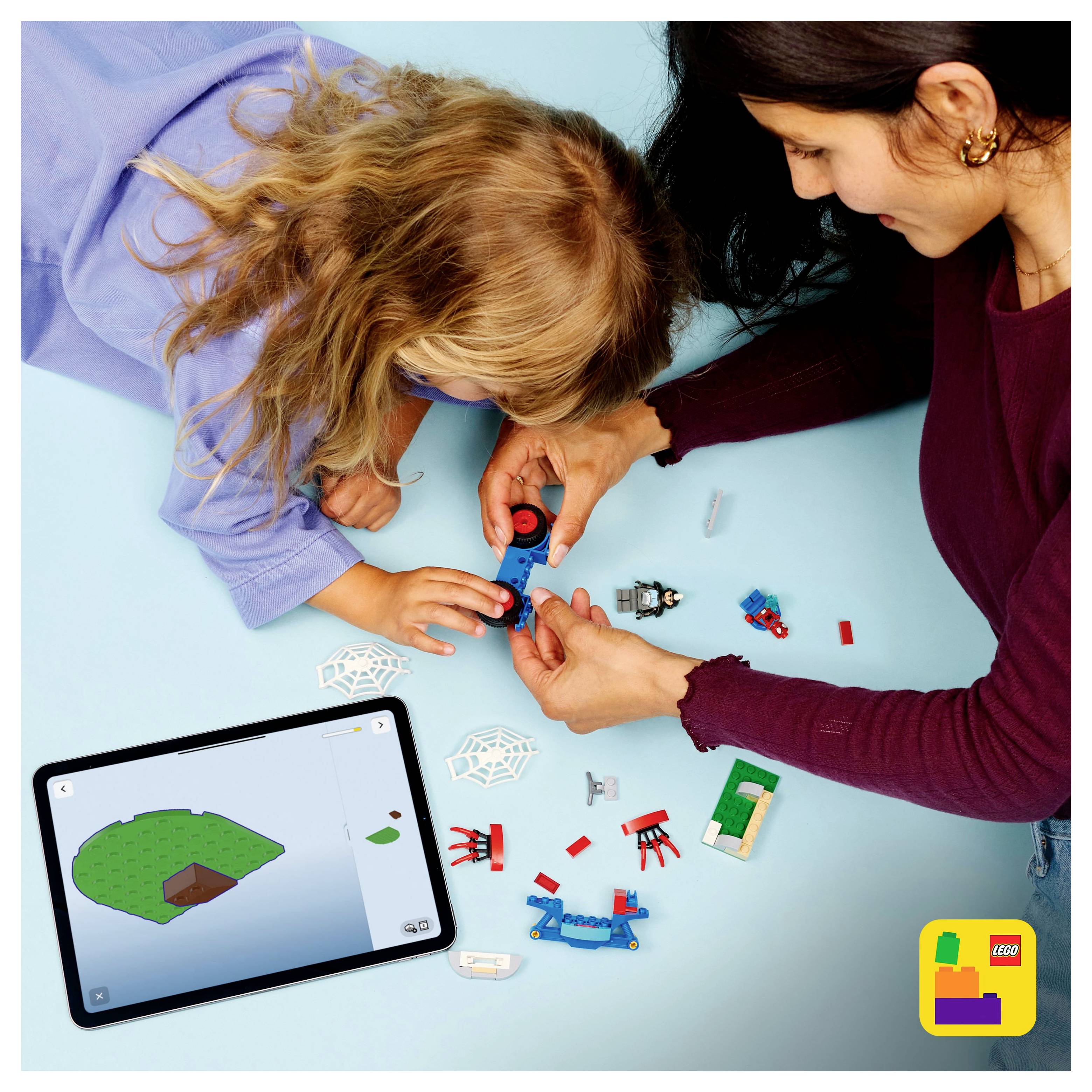 Ein Kind und ein Erwachsener bauen ein buntes Modell mit kleinen Bausteinen auf einer blauen Oberfläche und folgen dabei den Anweisungen, die auf einem Tablet angezeigt werden.