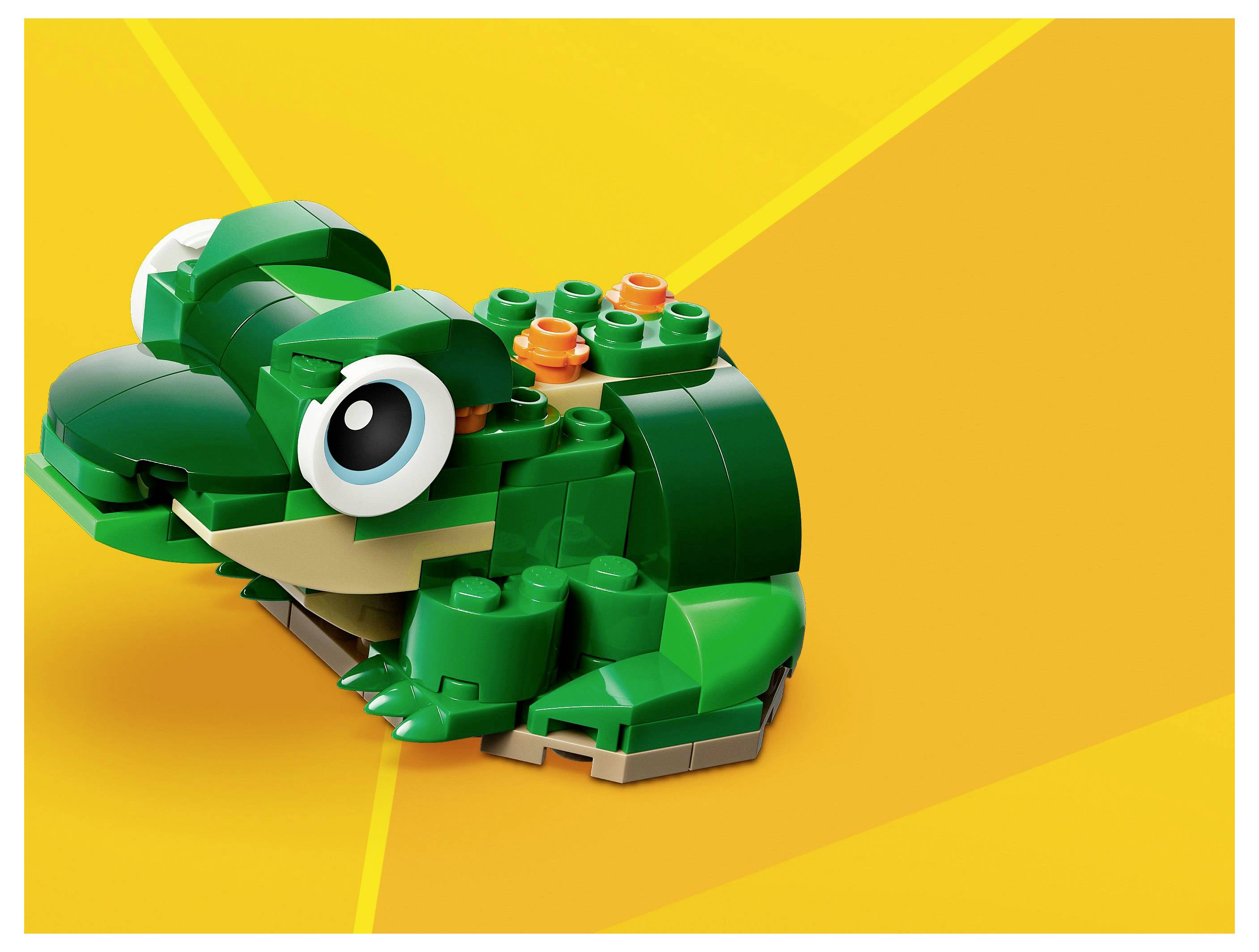 Ein grüner Spielzeugfrosch aus Bausteinen auf orangefarbenem Hintergrund, mit großen, comichaften Augen und einem lächelnden Mund.