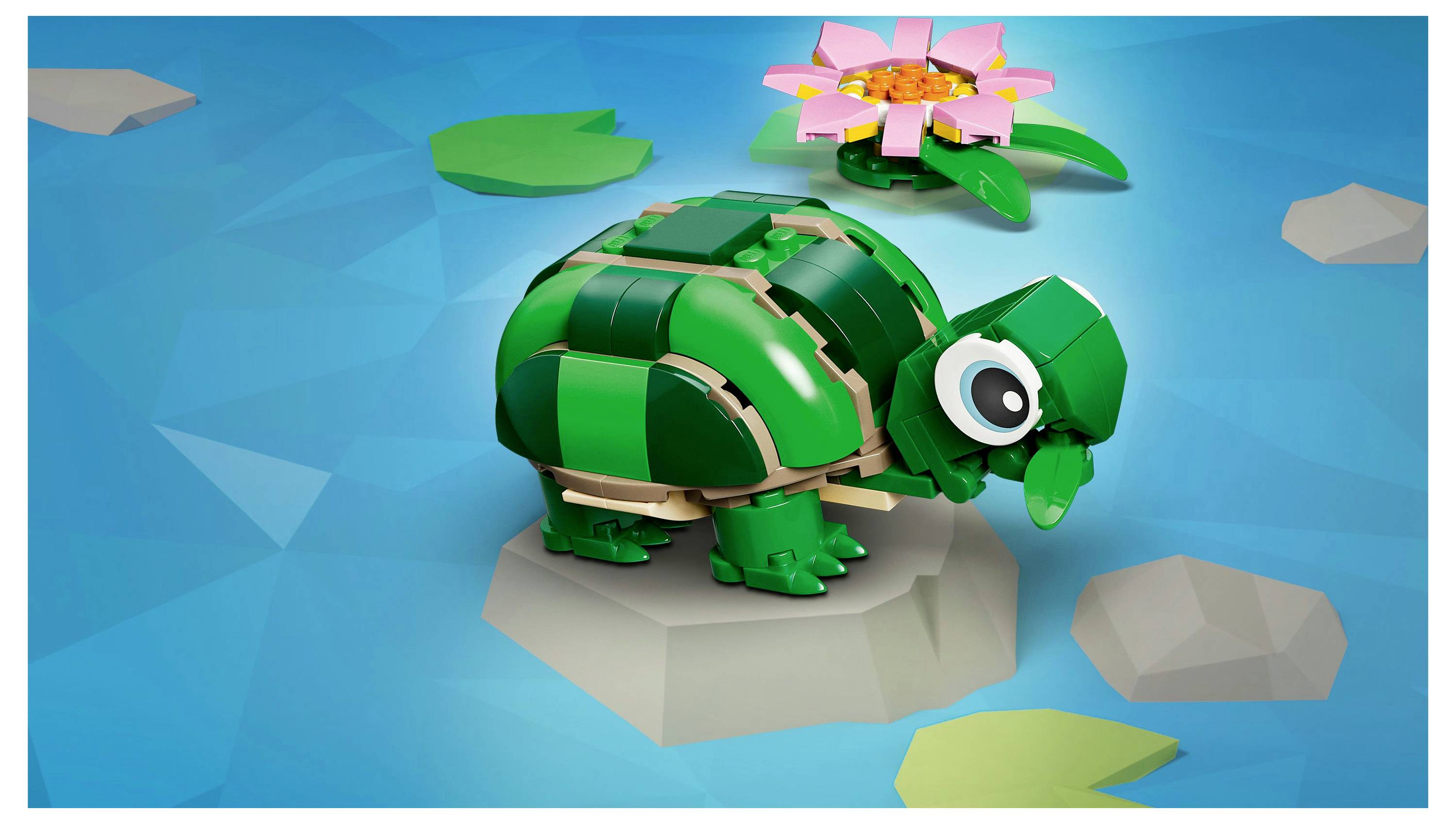 Eine kleine Schildkröte aus grünen Bauklötzen steht auf einem Stein im Wasser, mit einer pinken Blumenblock und verstreuten grünen und grauen Stücken drumherum.