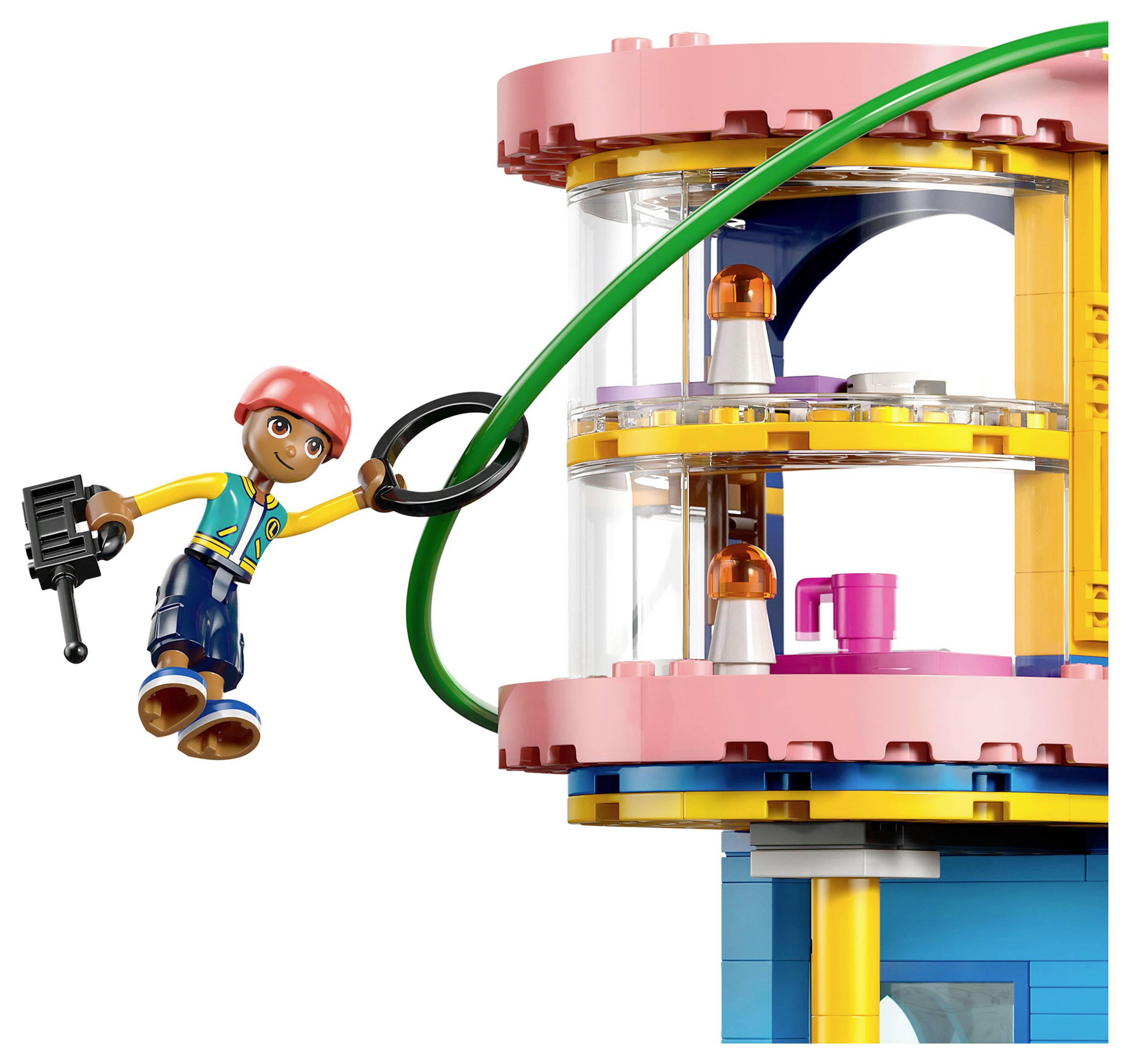 Eine LEGO-Figur mit einem roten Helm schwingt an einer grünen Liane in der Nähe eines mehrfarbigen Gebäudes mit zwei Etagen und großen Fenstern, in denen Möbel zu sehen sind.