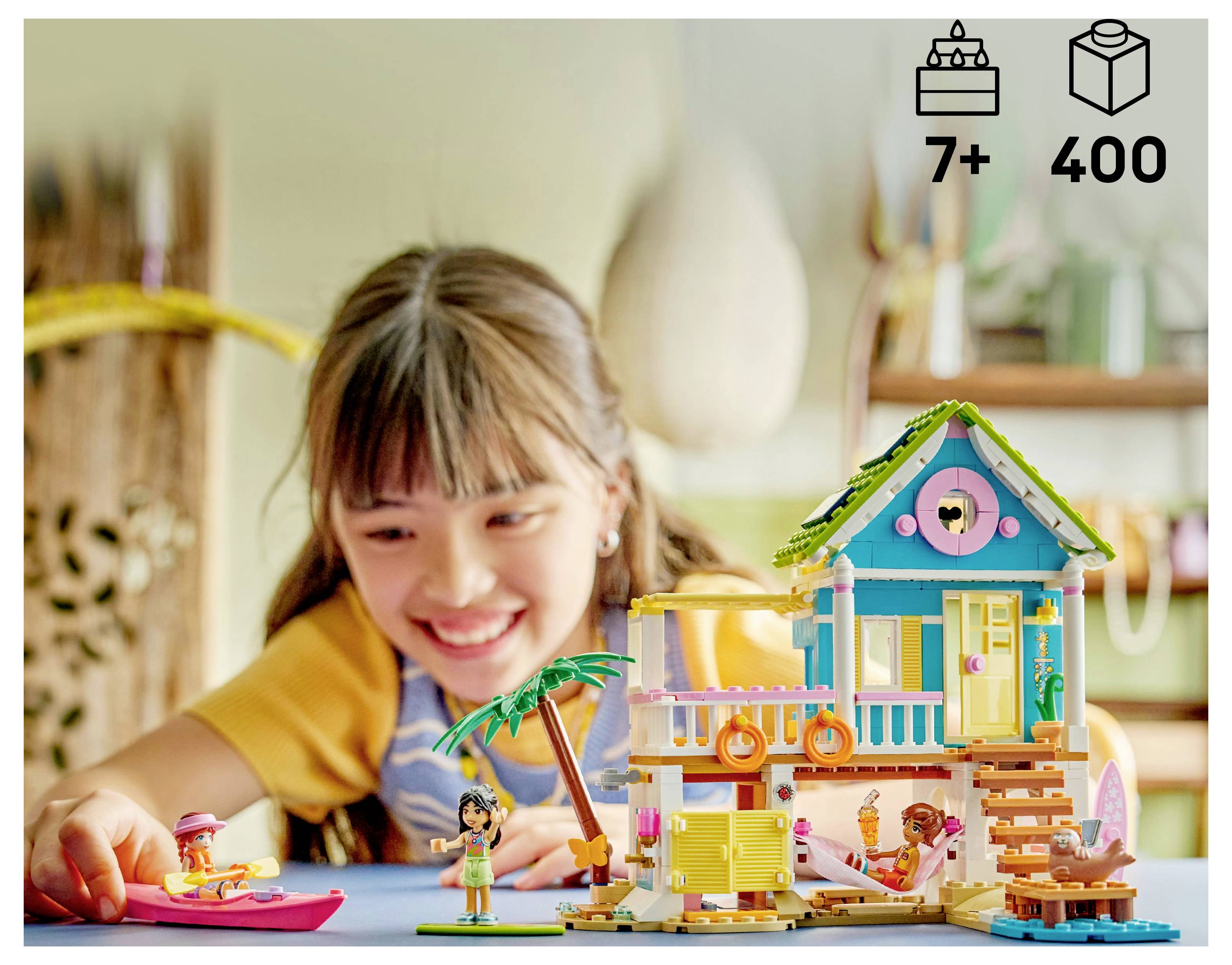 Ein Kind spielt mit einem bunten Strand-Spielset, das ein Haus, eine Palme und kleine Figuren enthält. Symbole zeigen das Alter 7+ und 400 Teile an.