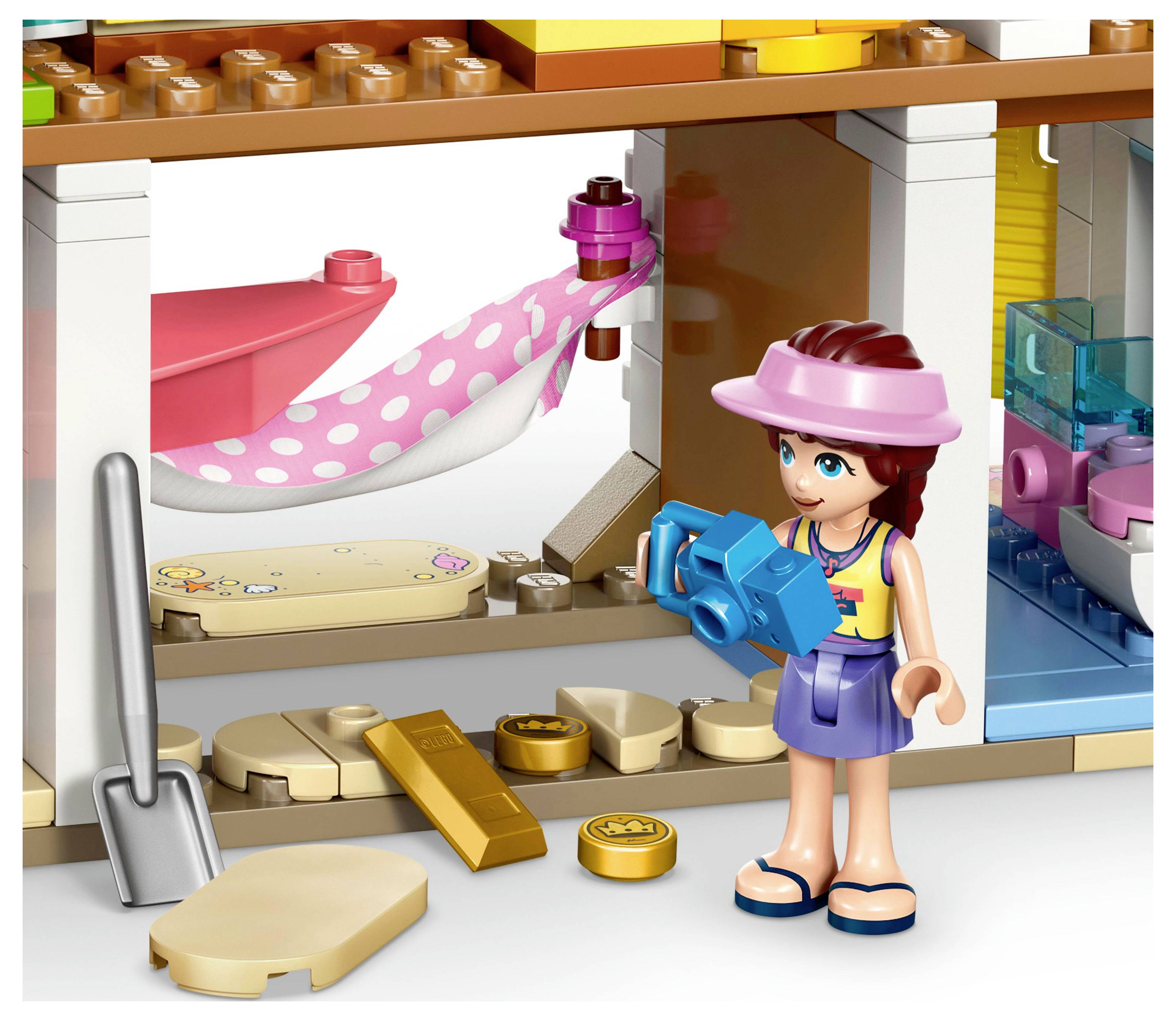 Eine Spielfigur mit einer Kamera steht in einem bunten Lego-Haus. Es gibt eine rosa Hängematte, eine Schaufel und verstreute Spielzeugmünzen.