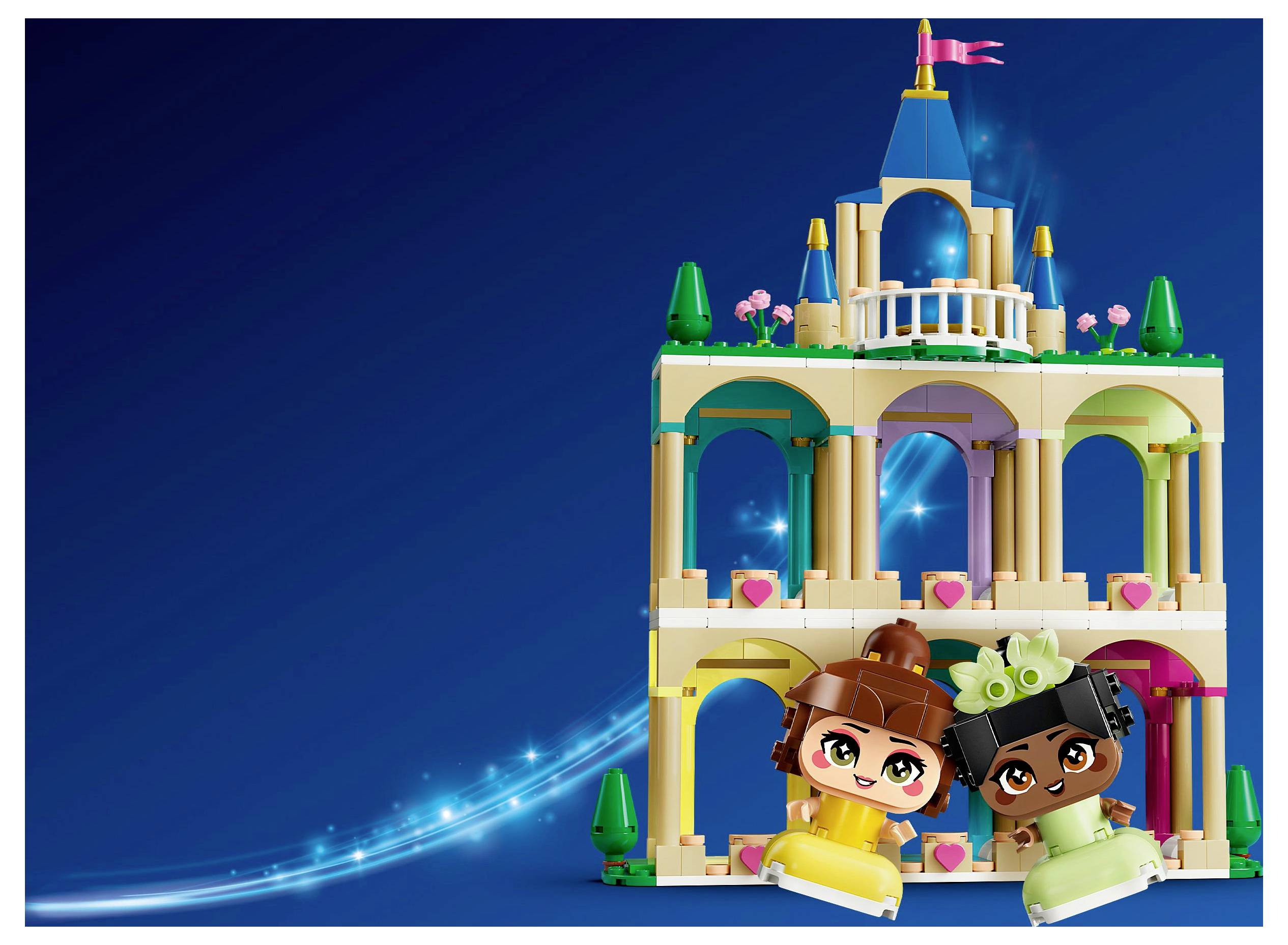 Zwei Spielfiguren in bunten Kostümen sitzen vor einem kleinen, kunstvoll verzierten Spielpalast mit Bögen und einem Turm, vor einem blauen Hintergrund.