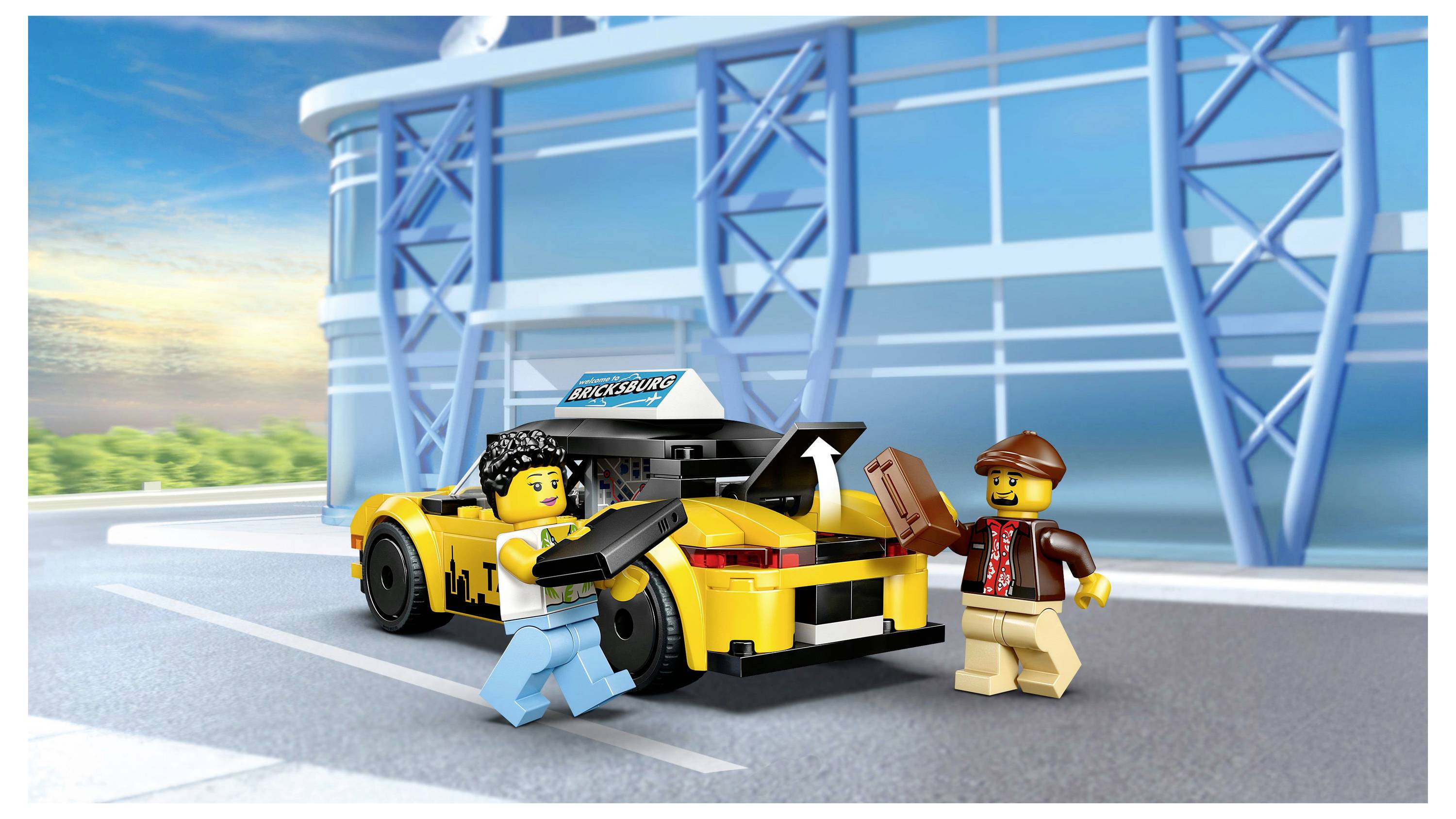 Zwei Spielfiguren mit Kameras stehen neben einem gelben Spielzeugauto vor einem Gebäude. Der blaue Himmel und die moderne Architektur bilden eine Kulisse.