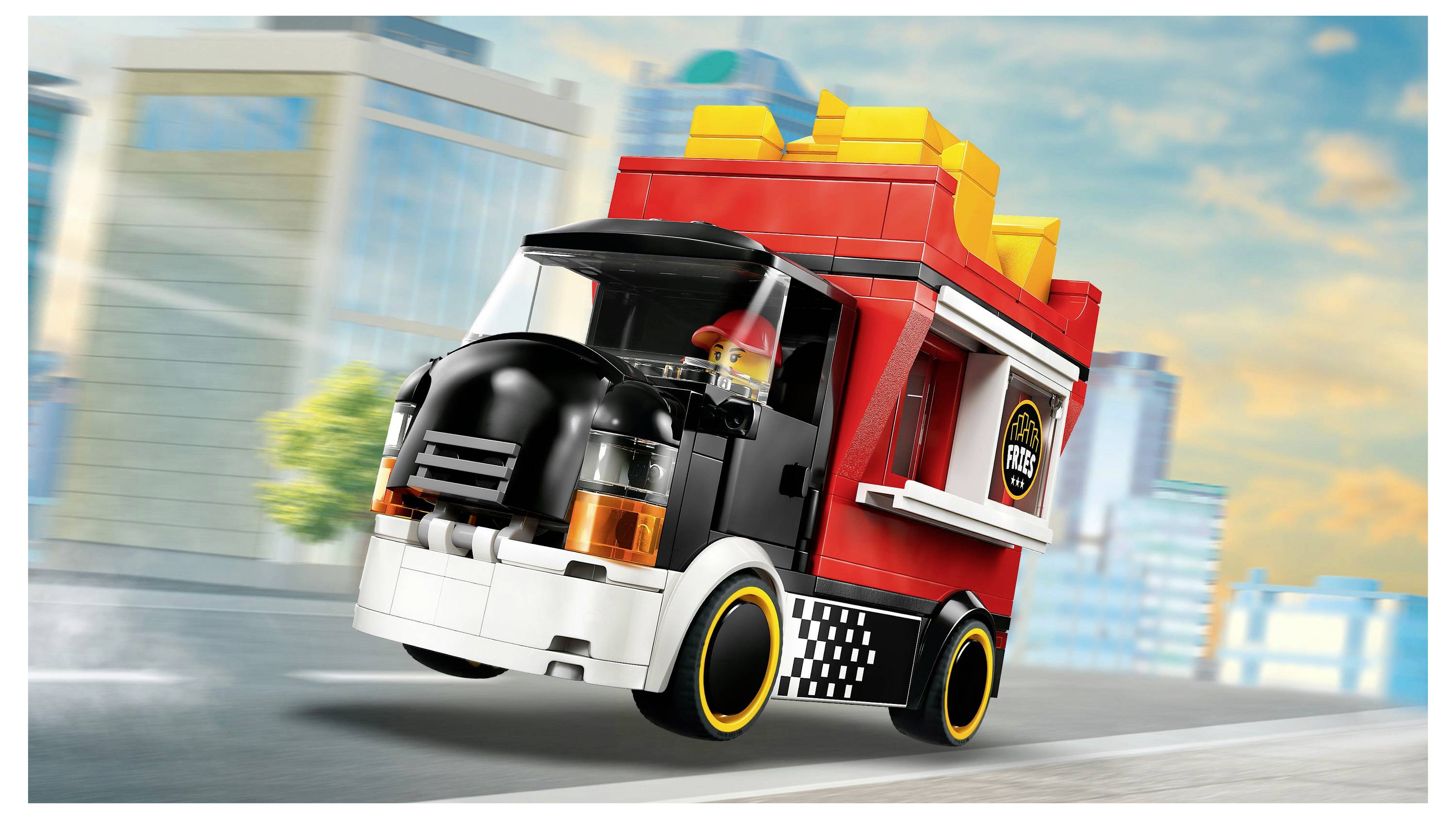 Ein rot-schwarzer Spielzeug-Lieferwagen, der aus ineinandergreifenden Plastiksteinen hergestellt wurde, fährt auf einer Stadtstraße mit Gebäuden im Hintergrund.