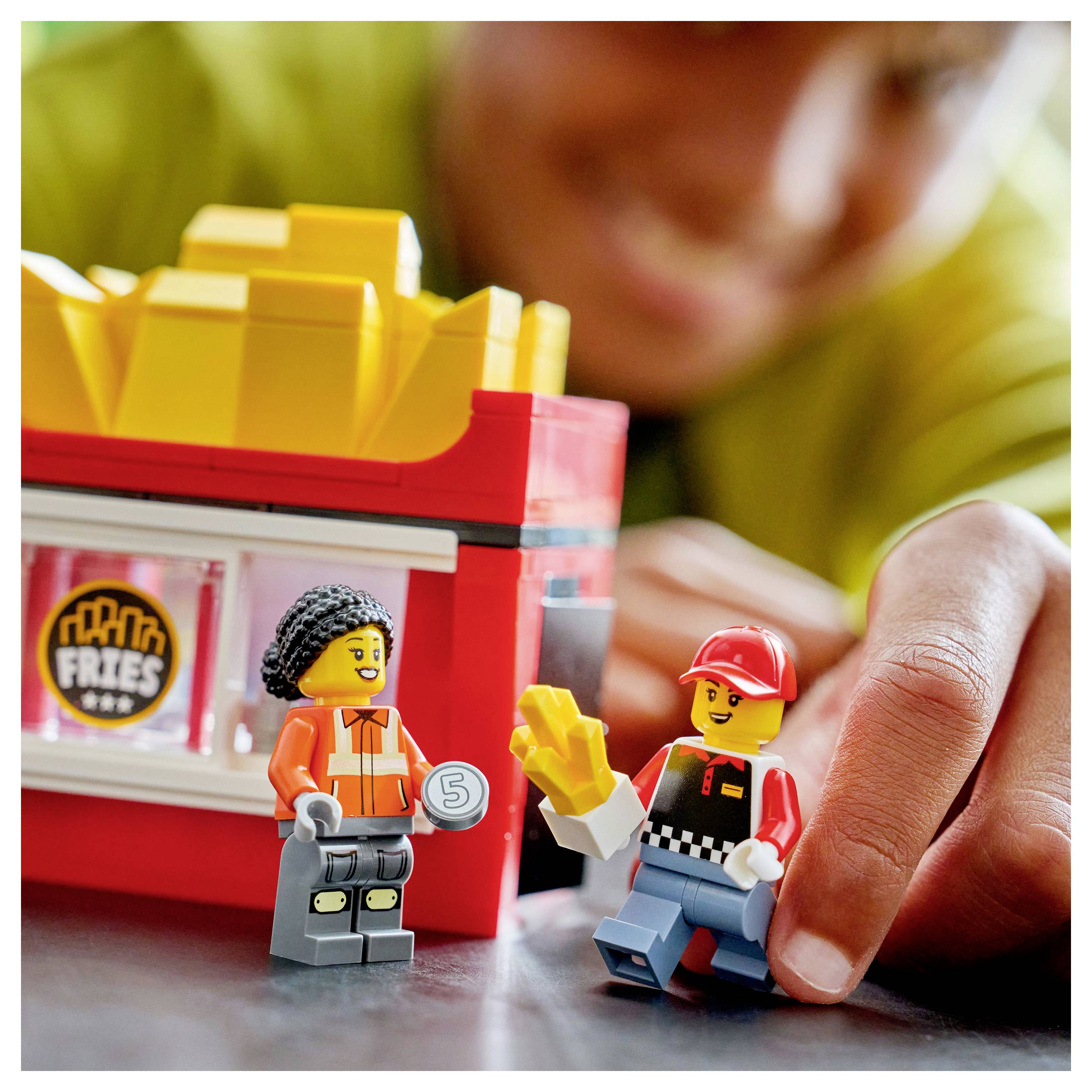 Ein Kind spielt mit Spielfiguren, die Arbeiter in einem Fast-Food-Umfeld nachbilden, wobei eine Figur Pommes hält und eine andere ein Objekt, das einem Geldzeichen ähnelt.