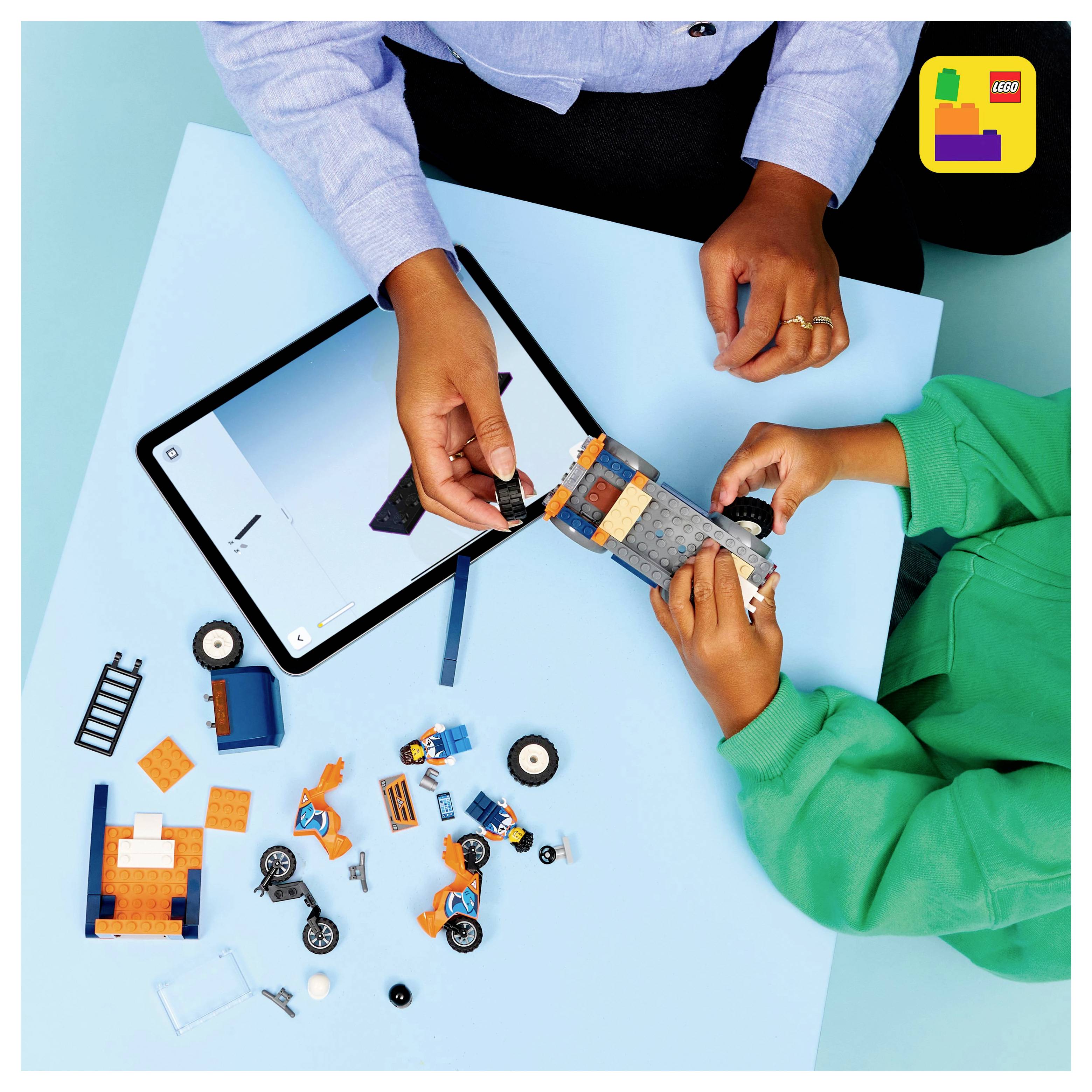 Zwei Personen bauen ein LEGO-Fahrzeug mit verschiedenen Teilen, neben einem Tablet mit Bauanleitungen. LEGO-Logo in der Ecke.
