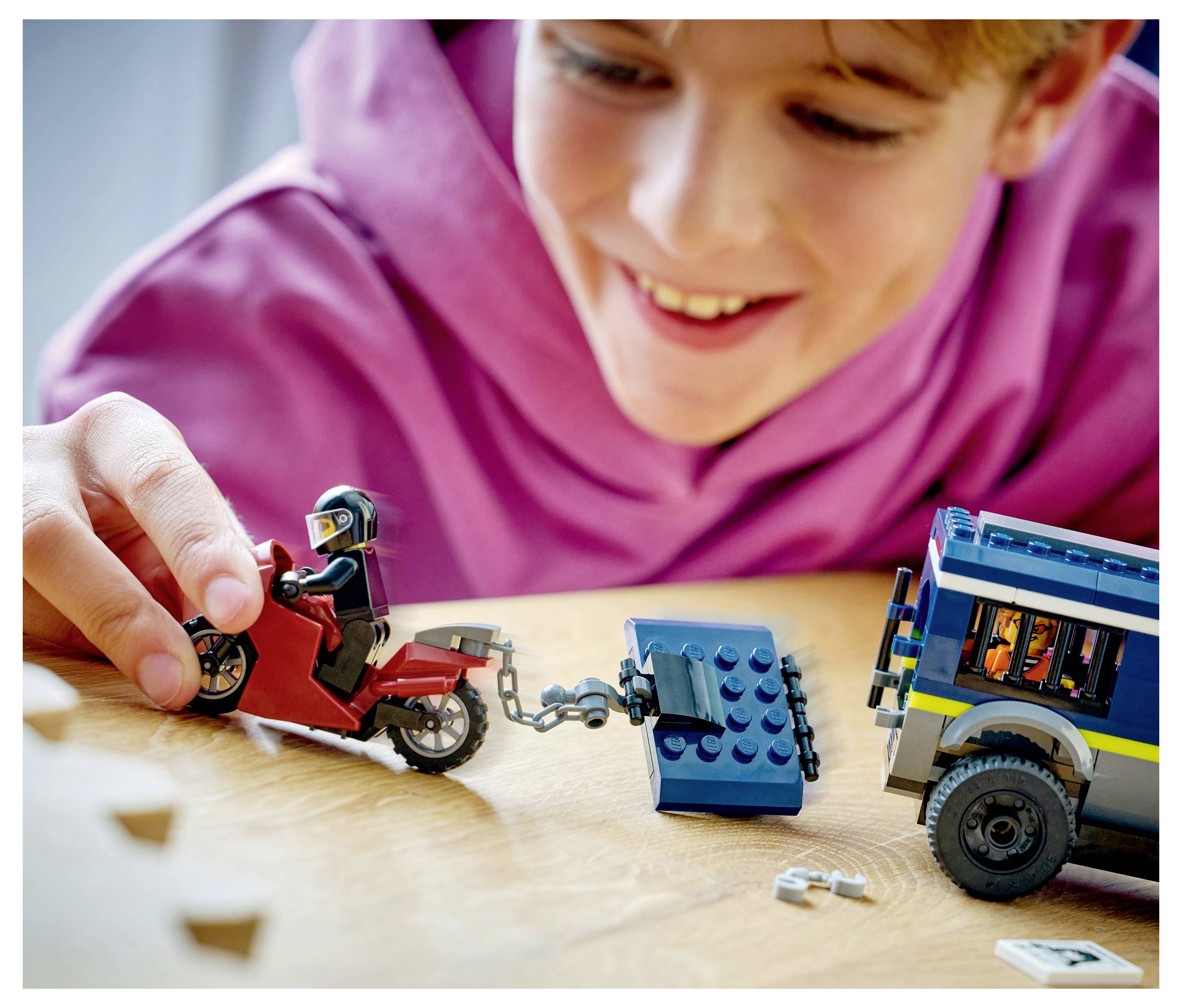 Ein Kind in einem pinken Hoodie spielt mit LEGO-Spielzeugen und bewegt ein rotes Motorrad und einen blauen Abschleppwagen auf einer Holzoberfläche.