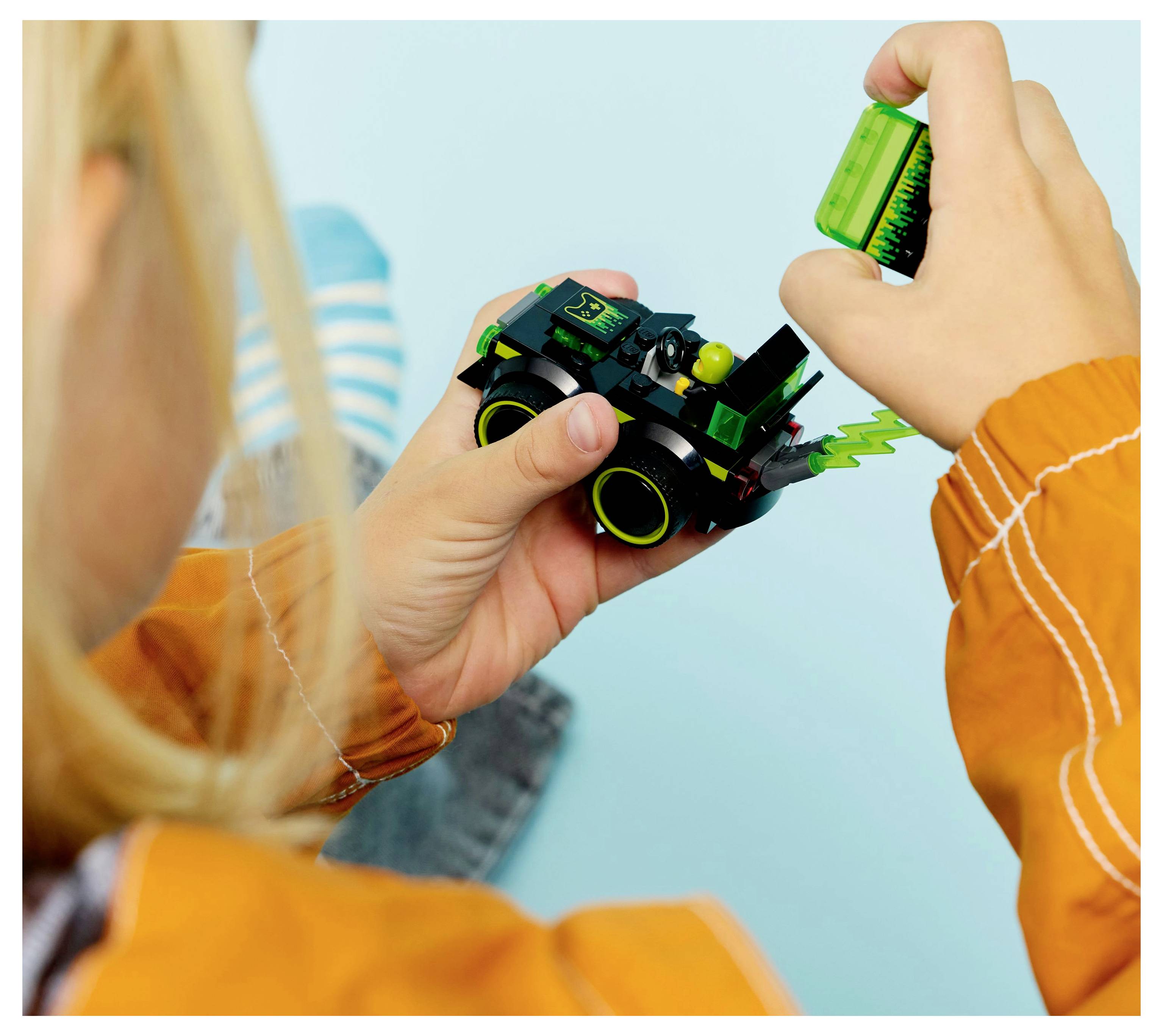 Ein Kind montiert ein kleines Spielzeugfahrzeug und konzentriert sich dabei mit äußerster Konzentration. Das Spielzeug ist grün mit gelben Rädern und hat ein Blitzentwurfsmuster. Die Ärmel des Kindes sind leuchtend orange.