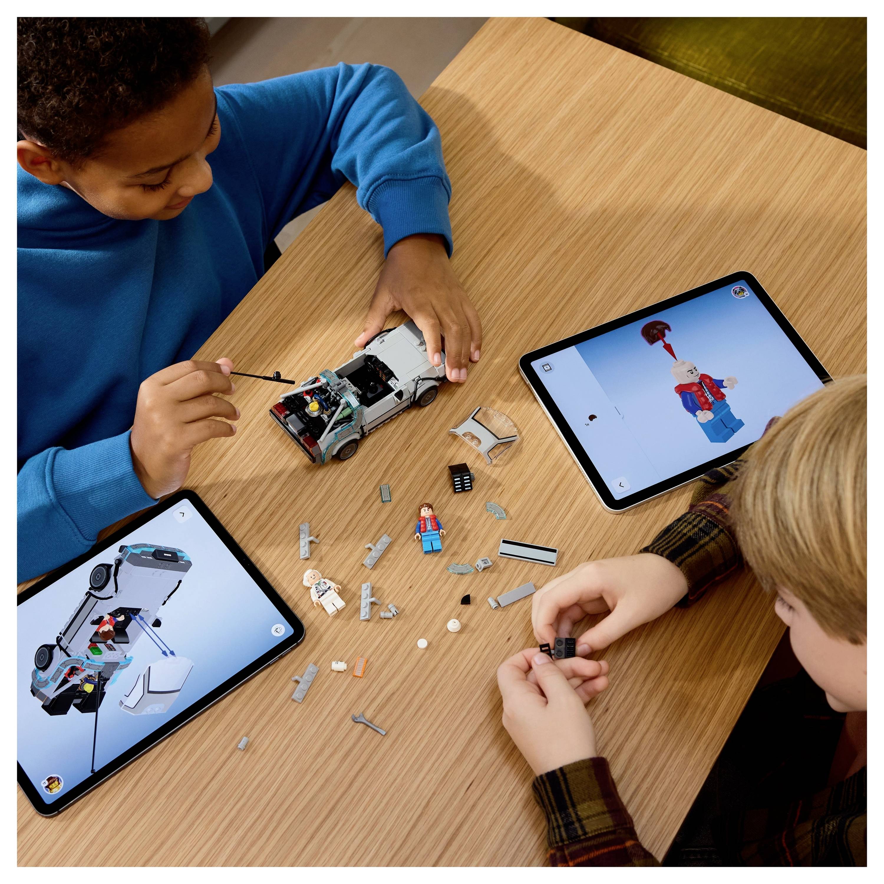 Zwei Kinder bauen ein Spielzeug-Modellauto mithilfe digitaler Anweisungen auf Tablets. Teile und ein teilweise zusammengebautes Automodell liegen auf dem Tisch zwischen ihnen.