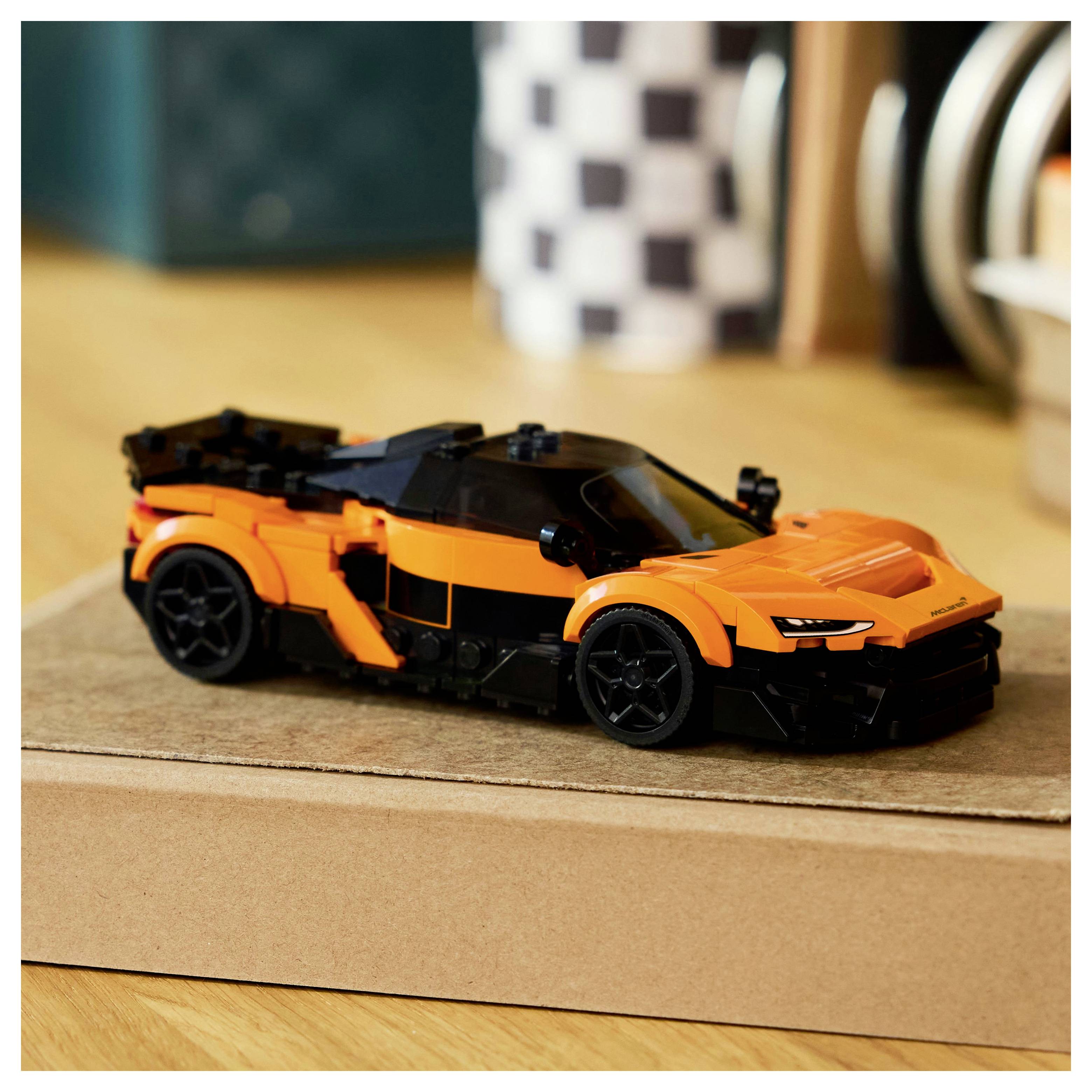 Ein kleines orangefarbenes Spielzeugauto mit schwarzen Details steht auf einer Pappschachtel, die auf einem Holzboden vor einem unscharfen Hintergrund platziert ist.