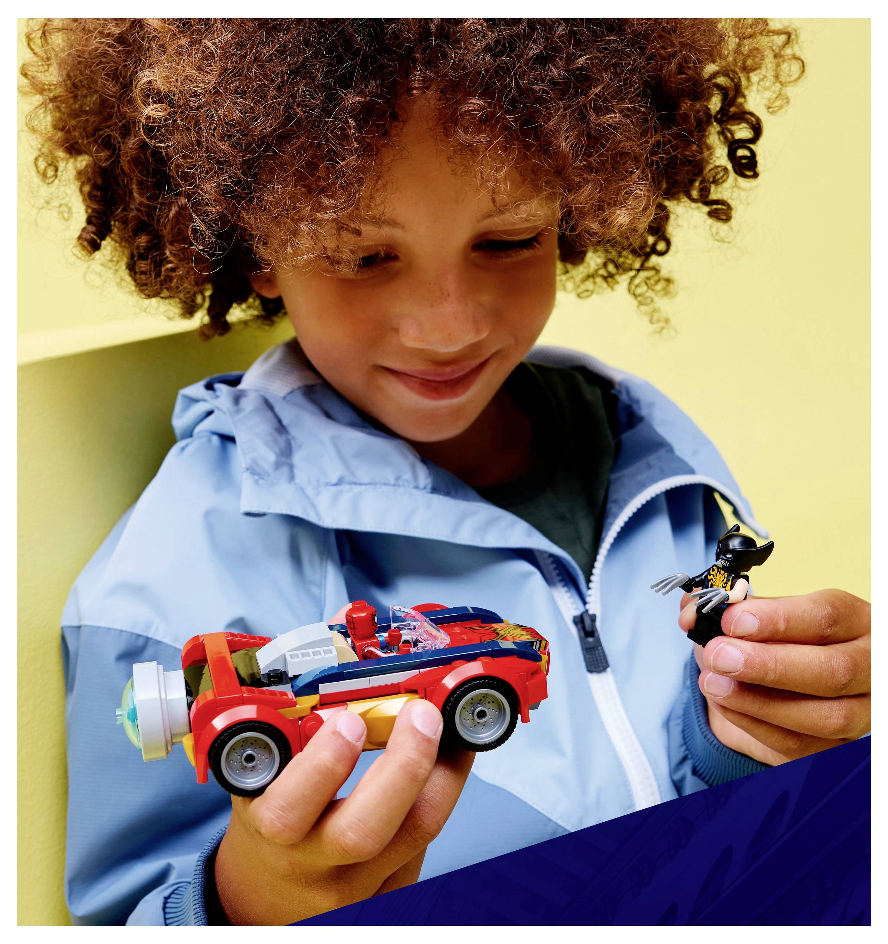 Ein Kind mit lockigem Haar, das eine blaue Jacke trägt, lächelt, während es einen roten Spielzeugwagen in der einen und eine Actionfigur in der anderen Hand hält, vor einem gelben Hintergrund.