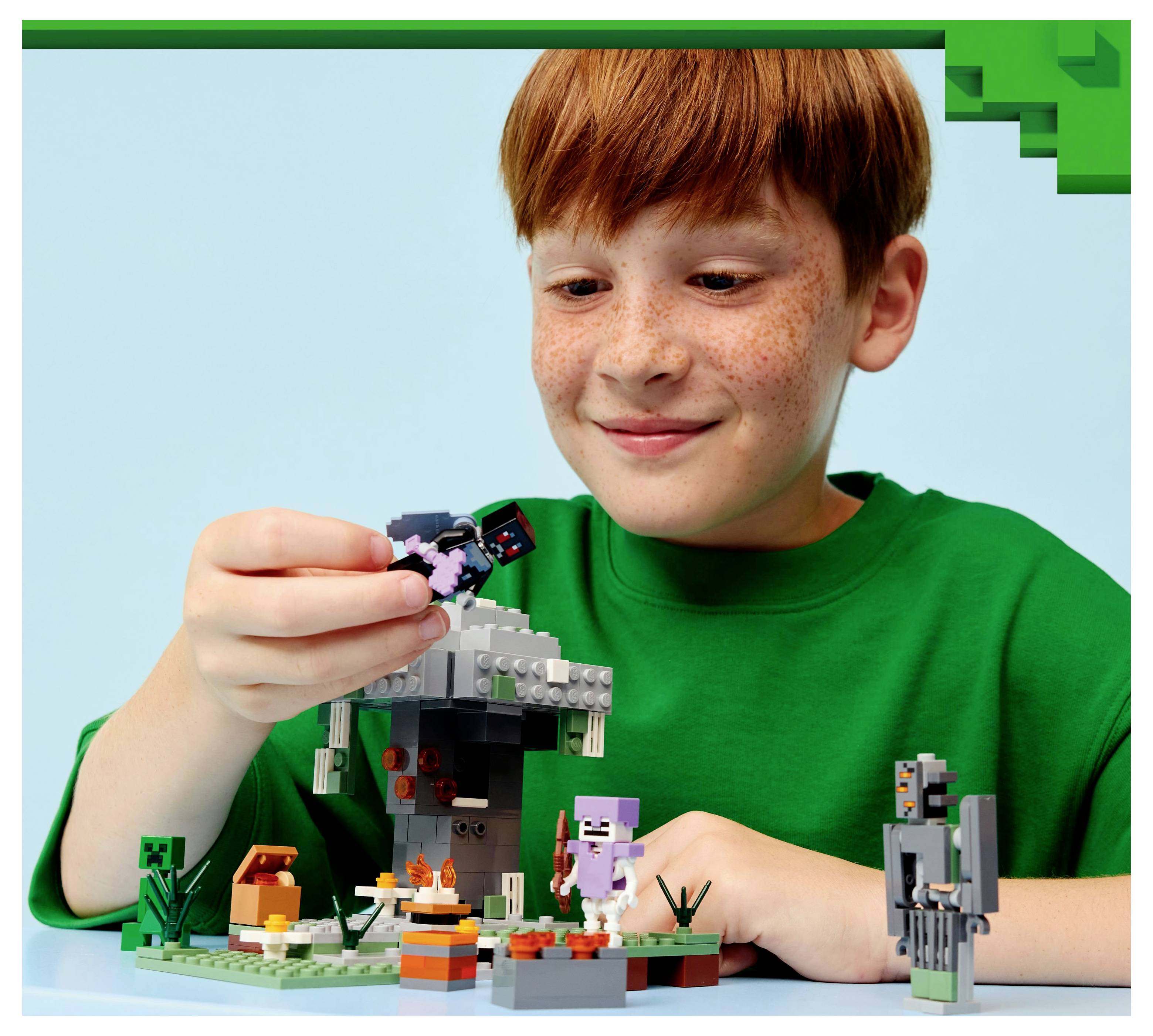 Ein Kind in einem grünen Shirt spielt mit einem LEGO-Set, das eine Minecraft-Szene mit kleinen Figuren und Zubehör auf einem Tisch zeigt.