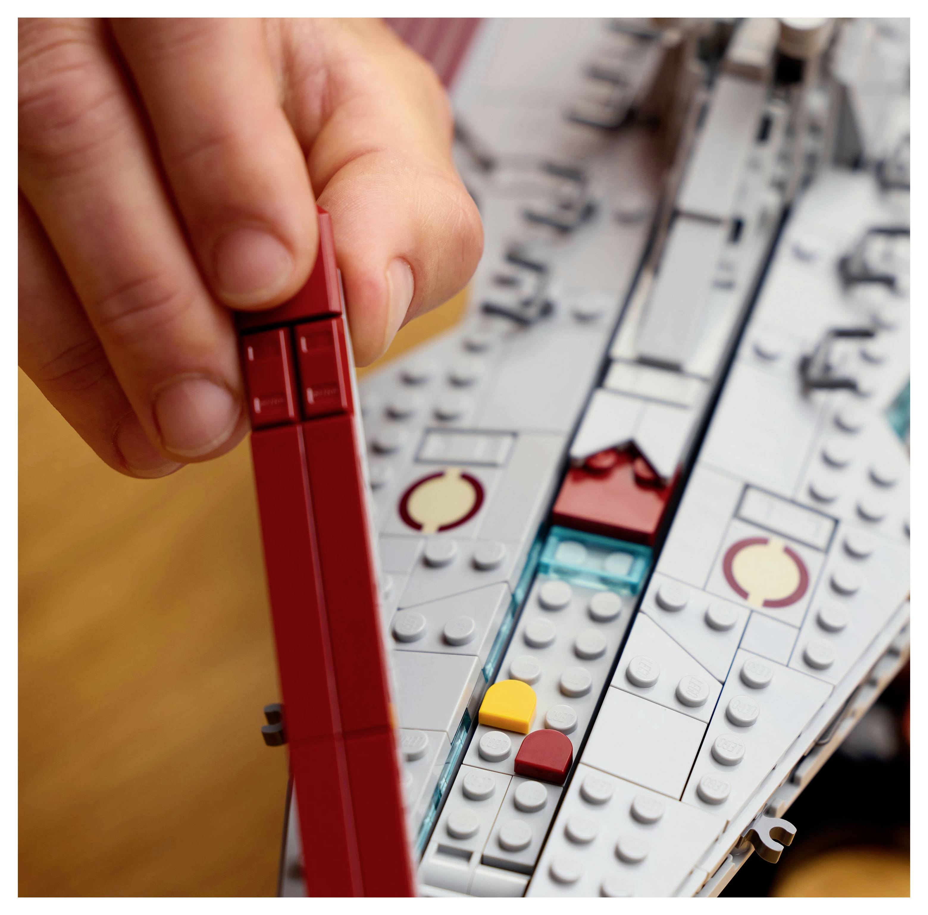 Eine Hand hält einen roten Stein über ein teilweise zusammengebautes Raumschiffmodell, das aus ineinandergreifenden Plastiksteinen konstruiert ist.