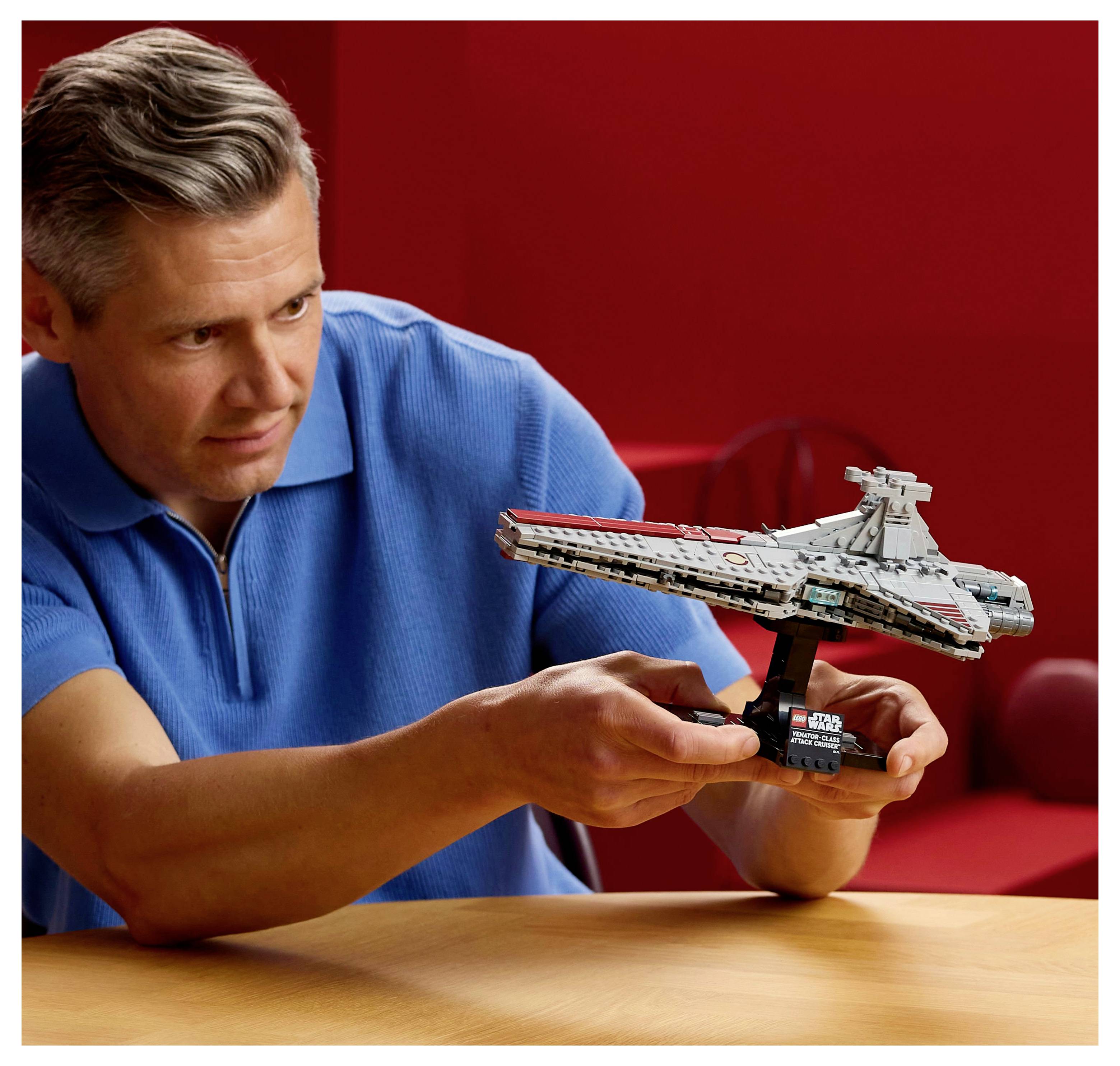 Eine Person in einem blauen Hemd hält ein detailliertes Modell eines Raumschiffs auf einem Ständer vor einem roten Hintergrund. Das Modell ähnelt einem fiktiven Raumfahrzeug.