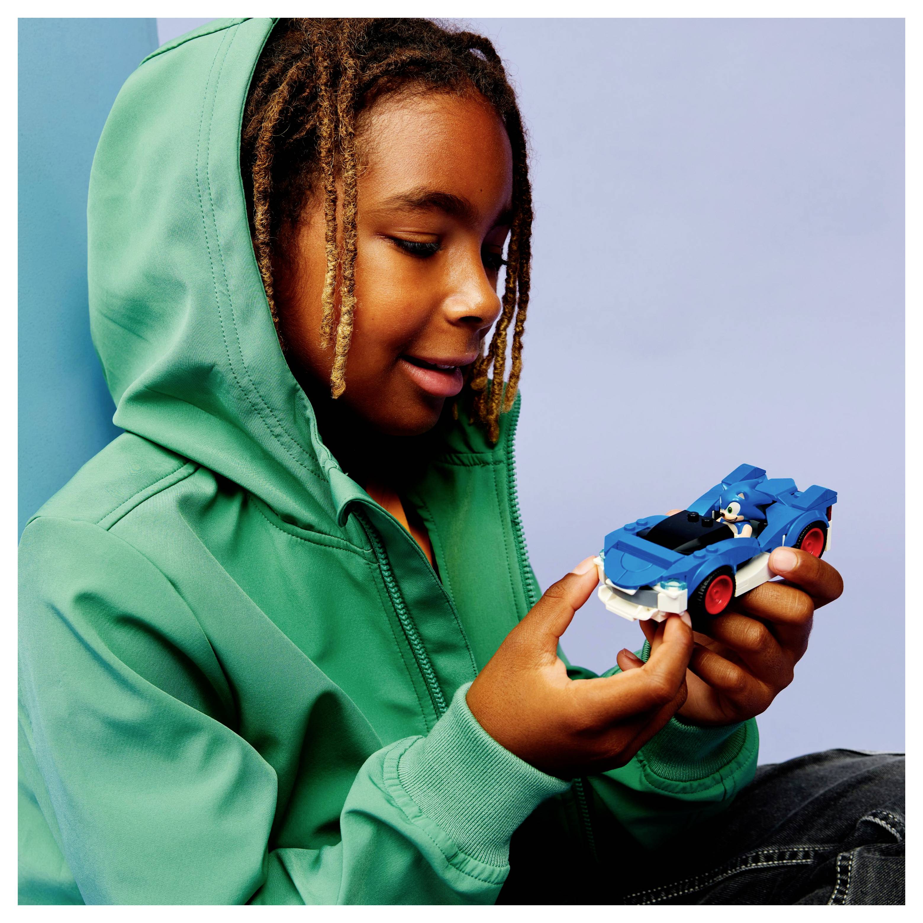 Ein Kind in einem grünen Hoodie lächelt, während es ein kleines blaues Spielzeugauto hält und vor einem hellblauen Hintergrund sitzt.