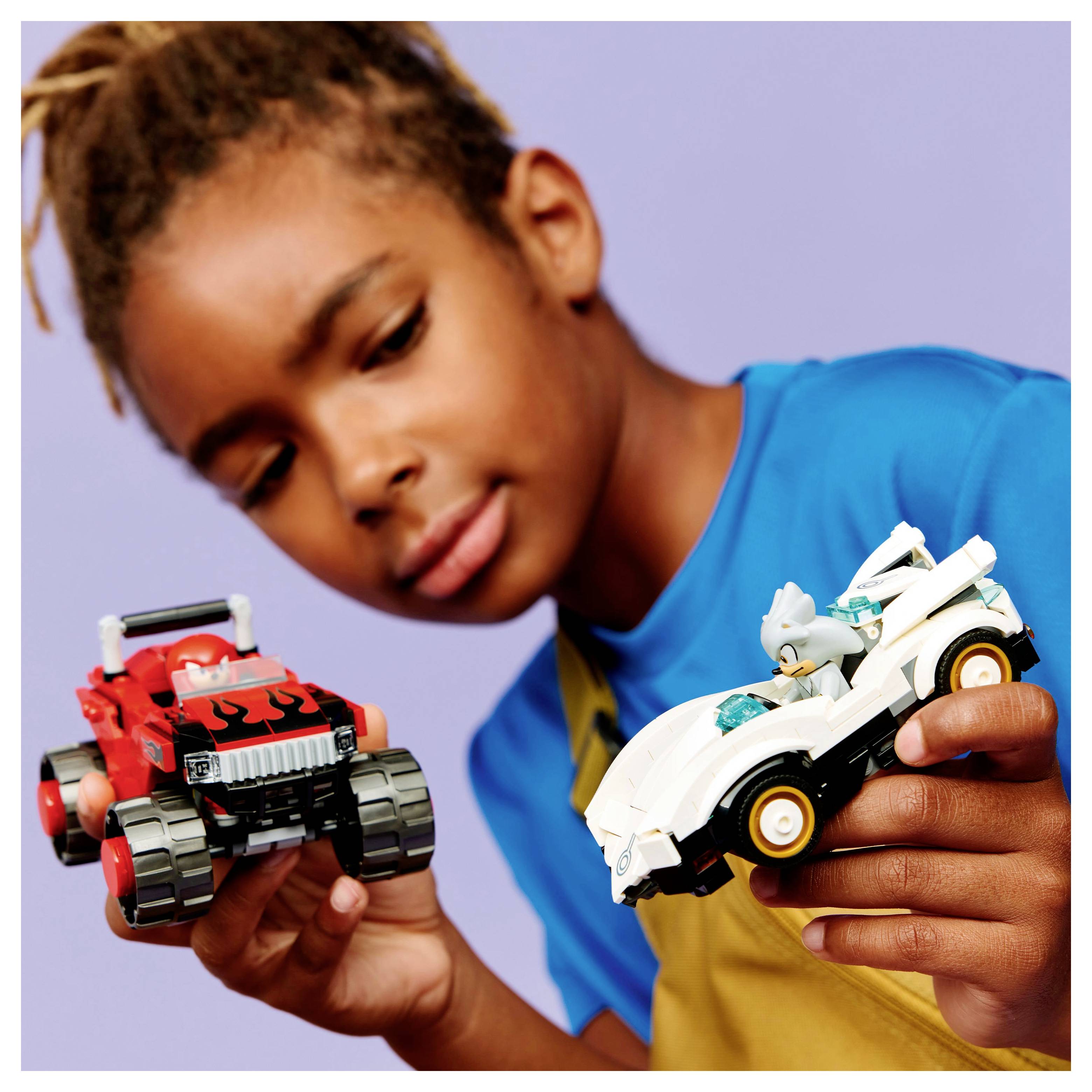 Ein Kind mit geflochtenen Haaren hält einen roten Spielzeugwagen in der einen Hand und einen weißen Spielzeugwagen mit einer kleinen Figur im Inneren in der anderen Hand.